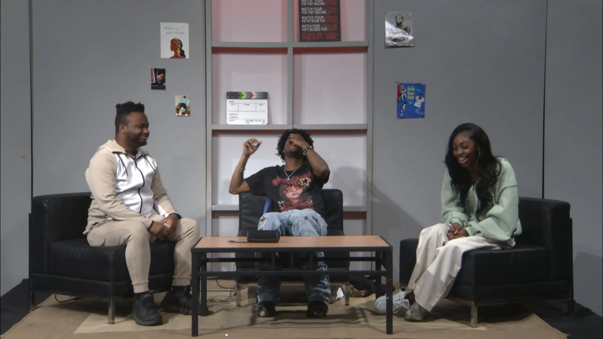 Three young adults sitting on black sofas in a casual room, laughing and smiling. The person in the middle is seated at a light wooden table, wearing a black t-shirt with a graphic, light blue jeans, and sneakers. The person on the left is dressed in a beige and white tracksuit with black boots, and the person on the right is dressed in a light green sweater and white pants. The background features gray walls decorated with posters and a shelf with a clapperboard and other items.