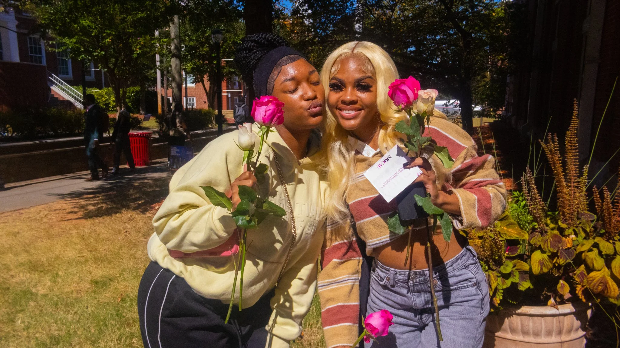 Two women standing close together outdoors, holding pink and white roses, smiling and touching cheeks. Background includes trees, flowers, and a brick building.