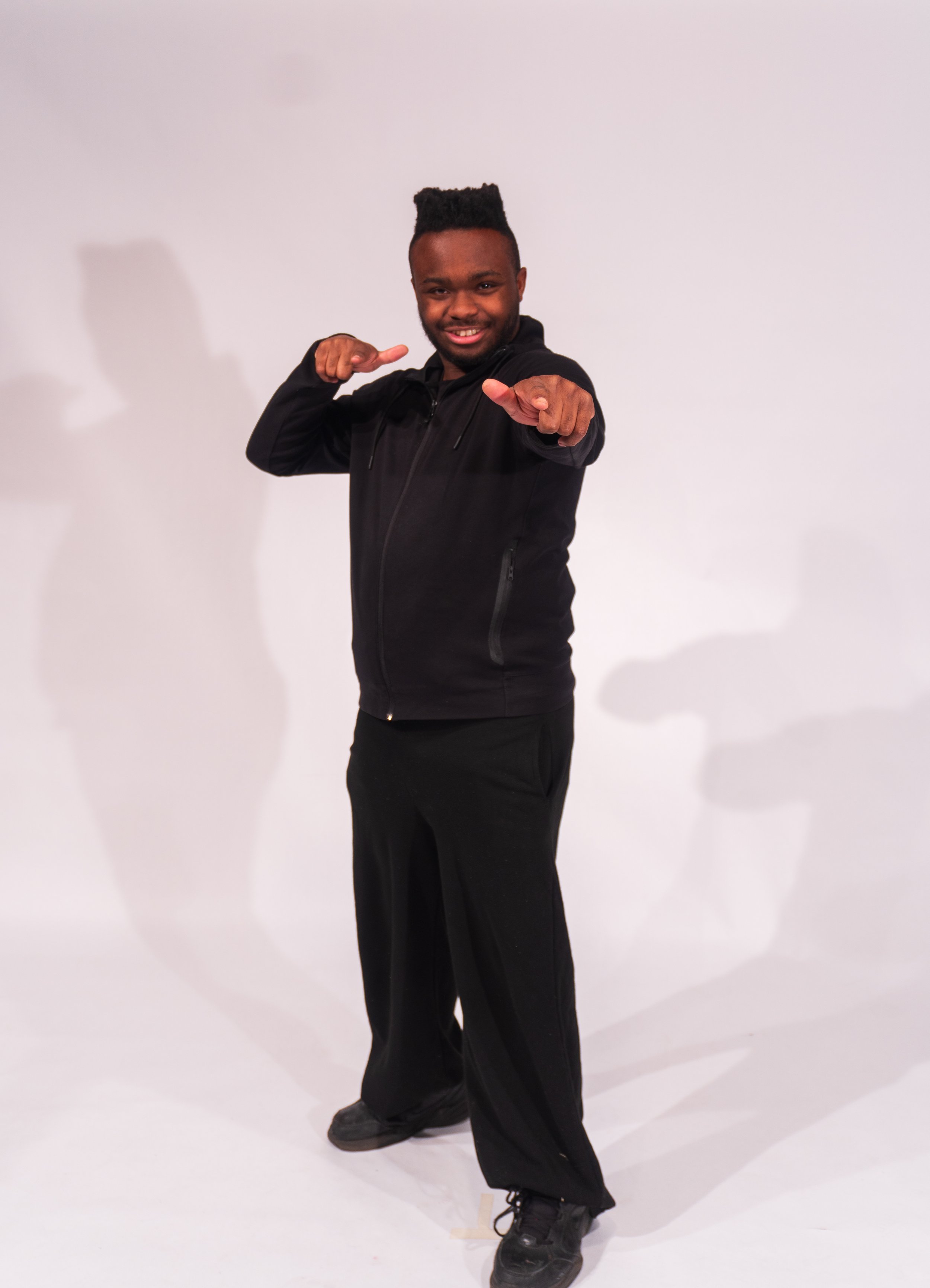 A young man with a tall flat-top hairstyle smiling and pointing towards the camera while standing against a plain white background.