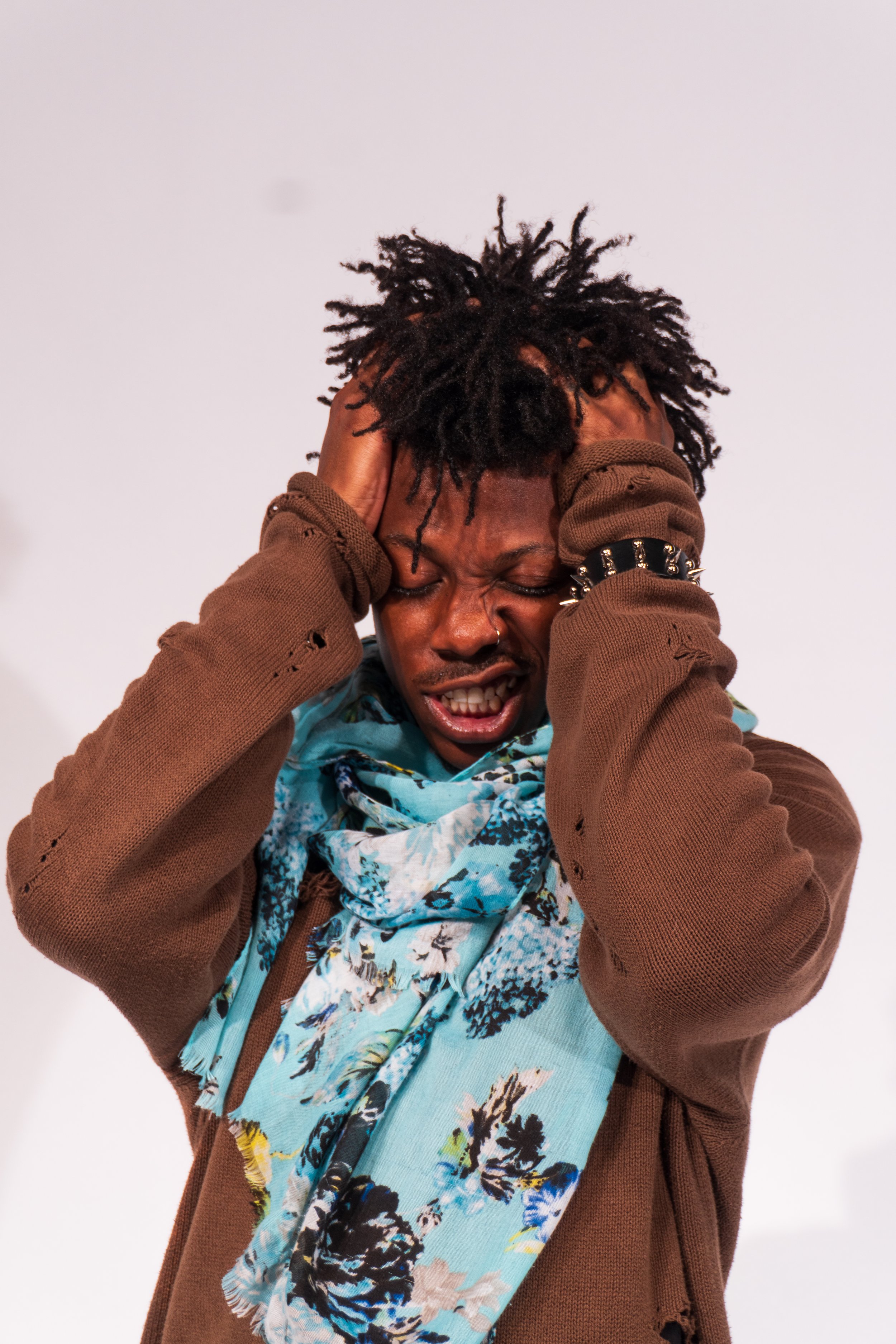 Person holding head with both hands, grimacing in distress, with closed eyes and mouth slightly open, wearing a brown distressed sweater and a floral scarf.