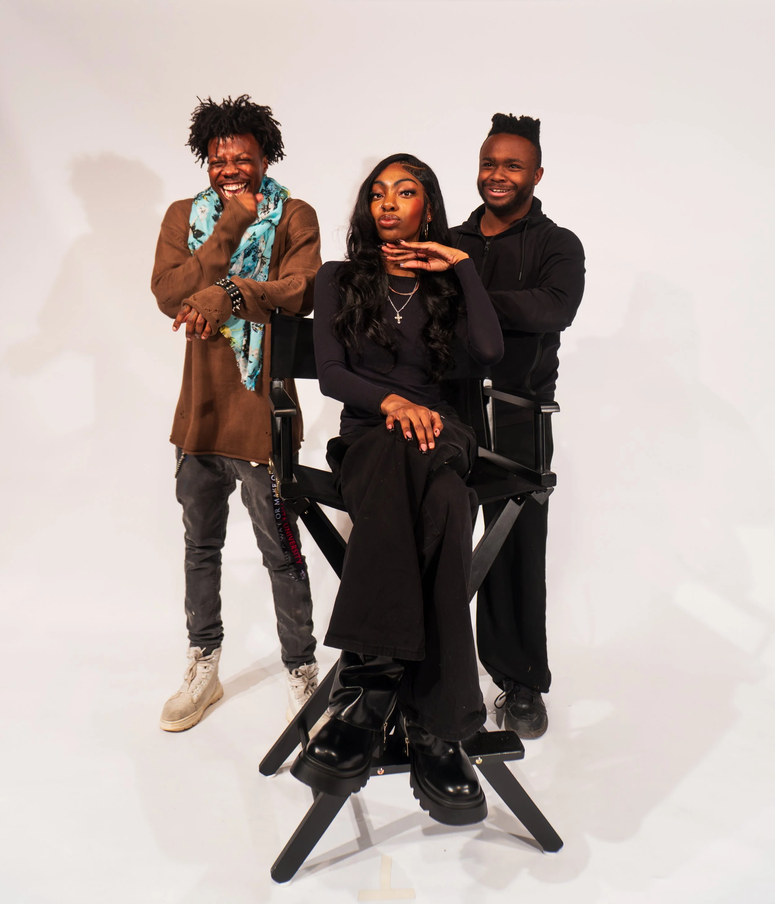 Three young adults posing for a photo against a plain white background. One woman seated on a director's chair, flanked by two men standing behind her. All are smiling and dressed casually, with the woman wearing black shoes and jewelry.