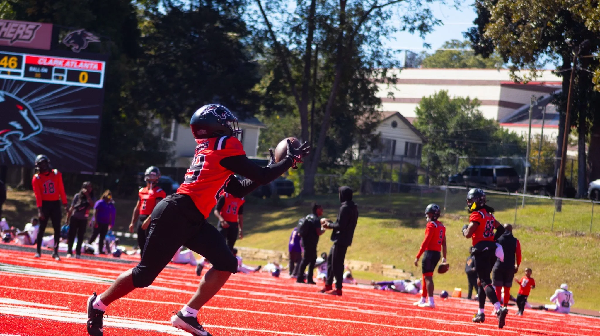 A football player wearing a black helmet, red and black jersey, and black pants catches a football on a red track field. Other players and coaches are in the background, with a scoreboard and trees surrounding the field.