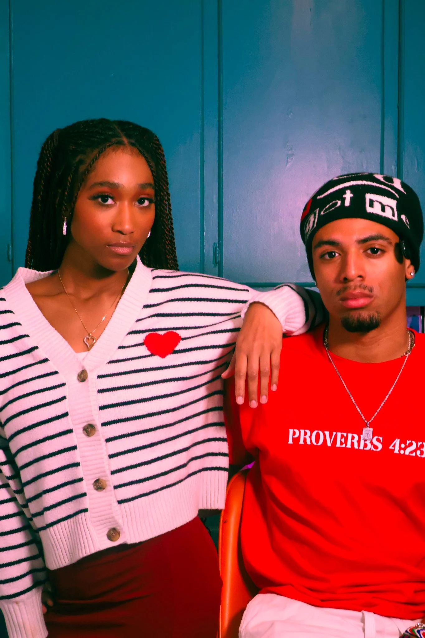 A young woman with braided hair and a young man wearing a beanie hat and red shirt seated together against a blue background, with the woman's arm resting on the man's shoulder.