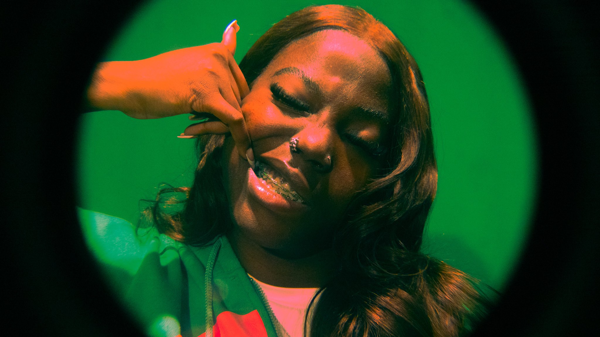 Close-up of a young woman with brown hair, wearing braces, making a gesture near her face, against a green background.