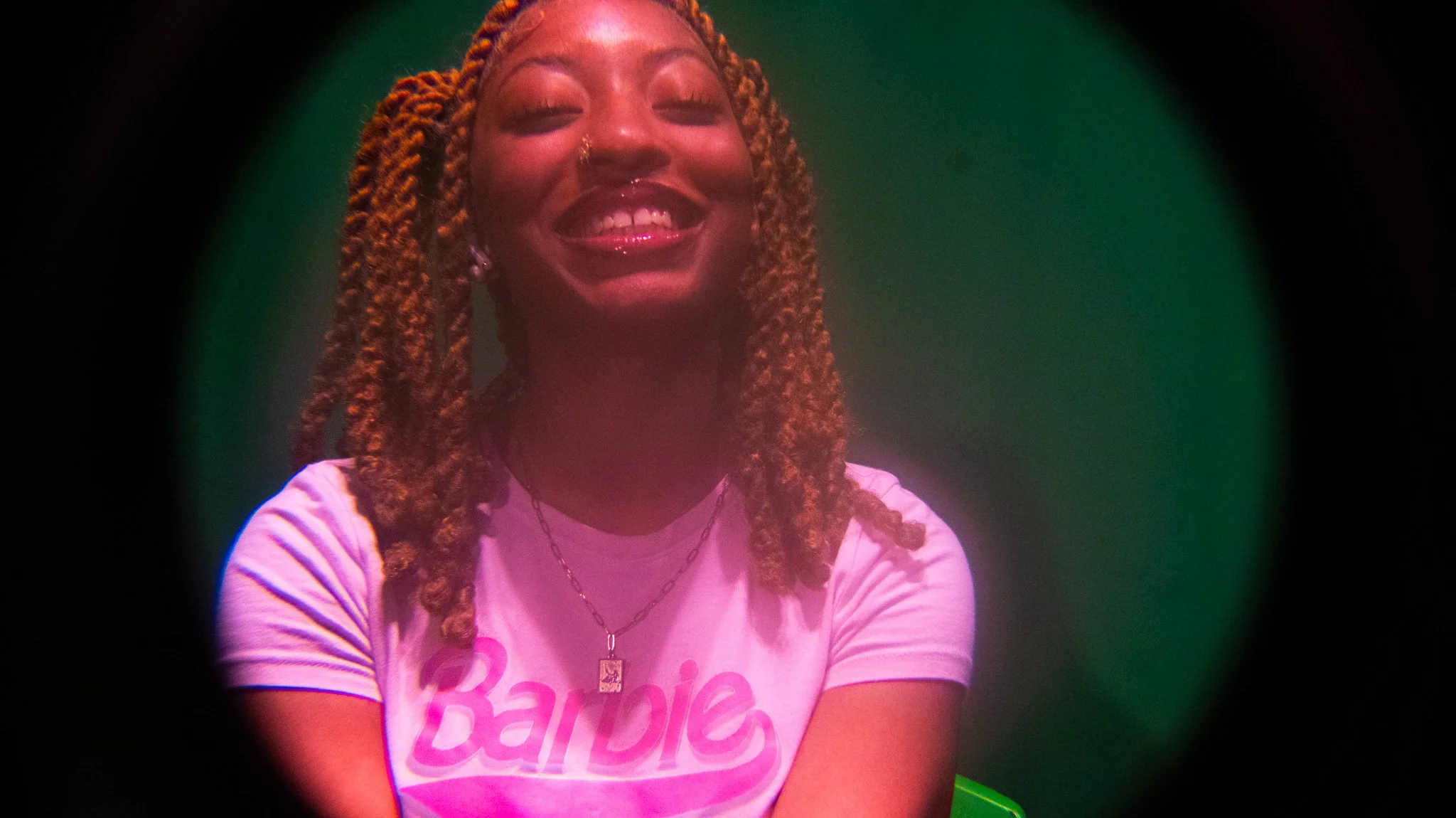A smiling woman with reddish-brown braids wearing a white t-shirt with pink 'Barbie' logo, necklace, and earrings, captured through a circular view with a green background.