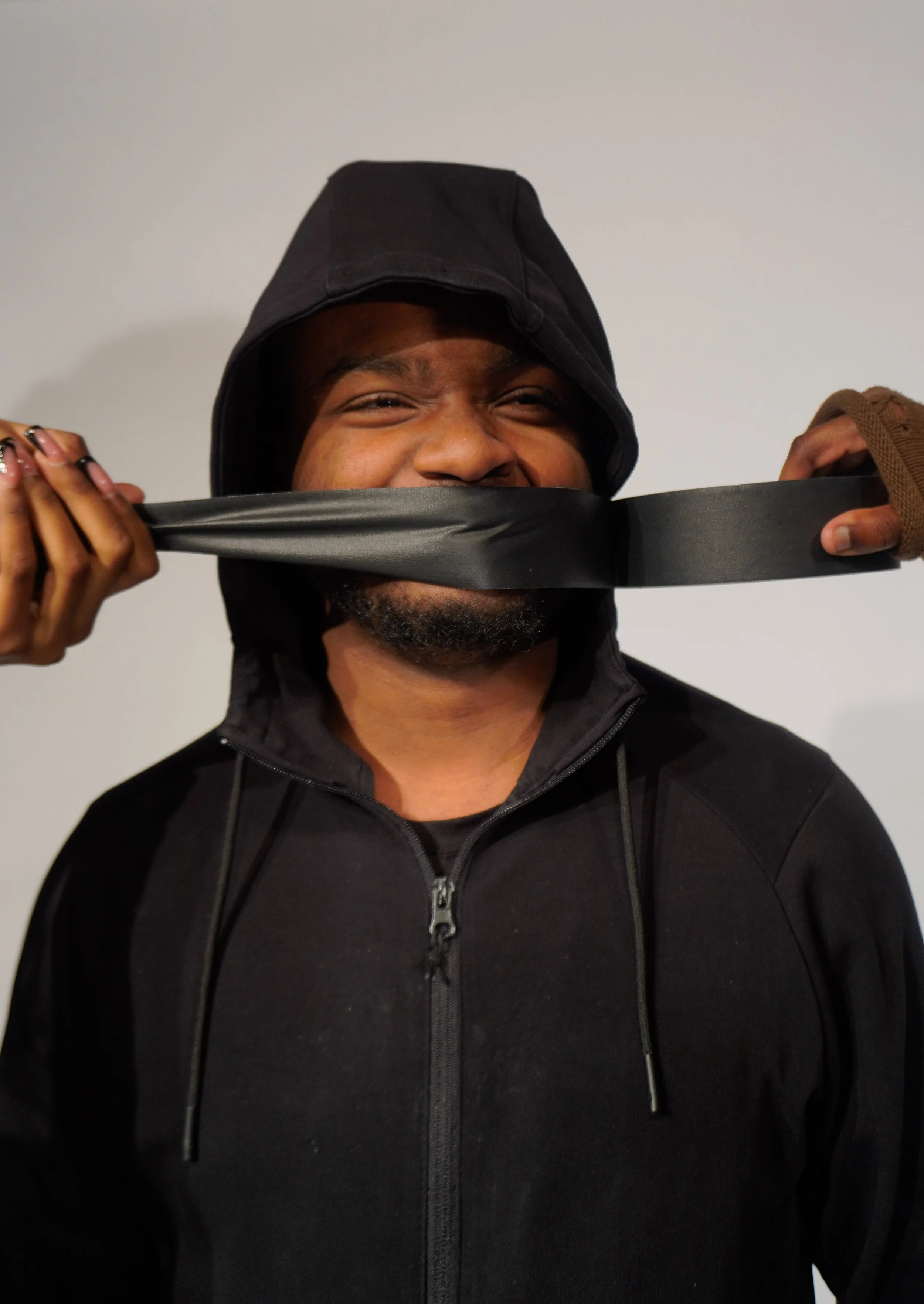 A person with a black hoodie being playfully gagged with a black strip of material, smiling with eyes slightly squinting.