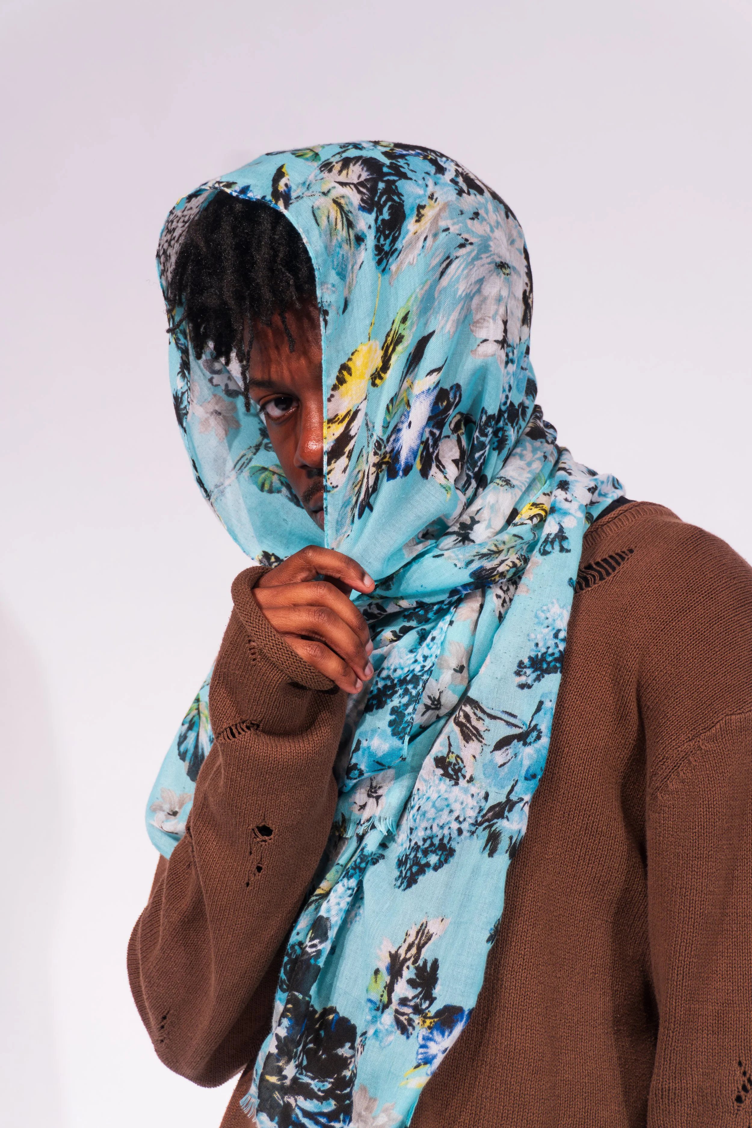 A person wearing a brown sweater and a colorful floral headscarf, partly covering their face, looking at the camera against a plain light background.