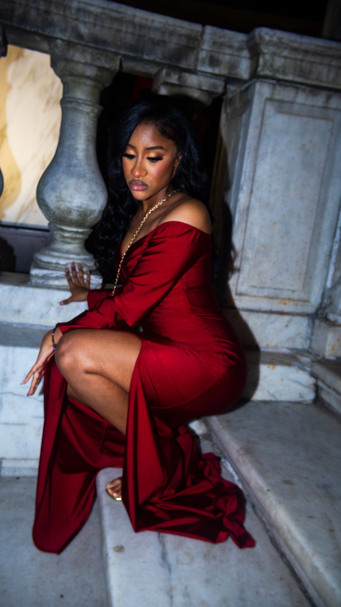 A woman in a long red off-shoulder dress kneeling on marble steps, leaning against a stone railing with columns, in a dimly lit setting.