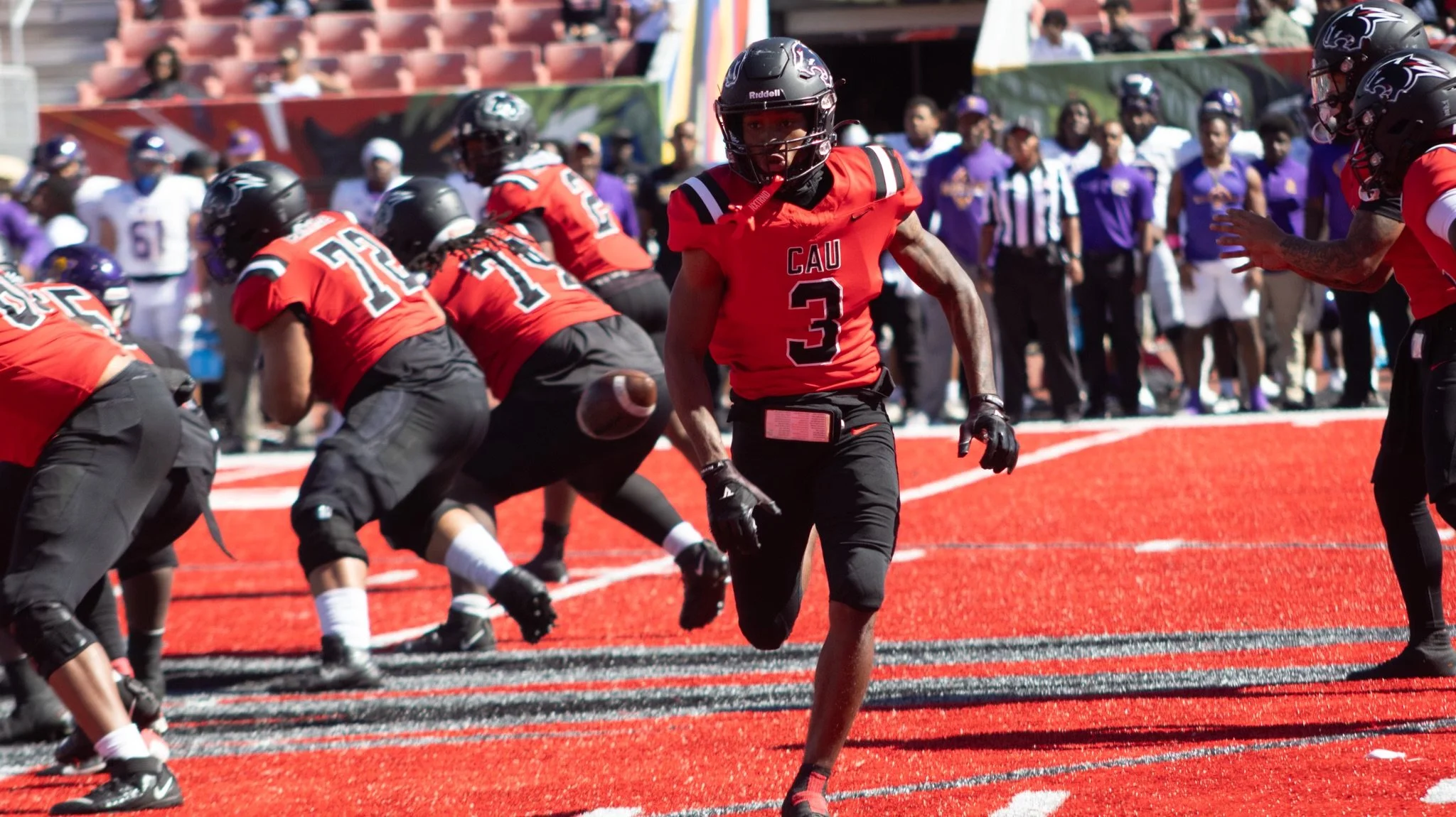 American football game with players in black helmets and red jerseys on a red field, with spectators in the background.