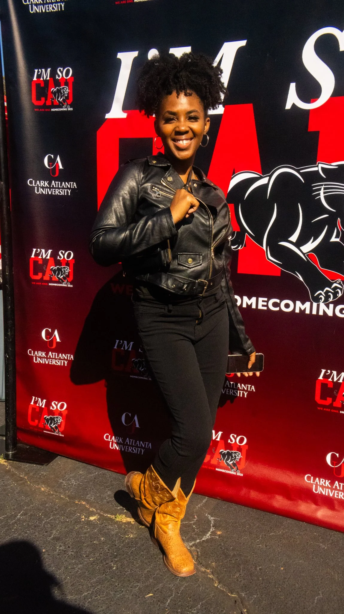 A woman smiling and posing in front of a Clark Atlanta University homecoming backdrop. She is wearing a black leather jacket, black jeans, and brown cowboy boots, holding a phone in her left hand.