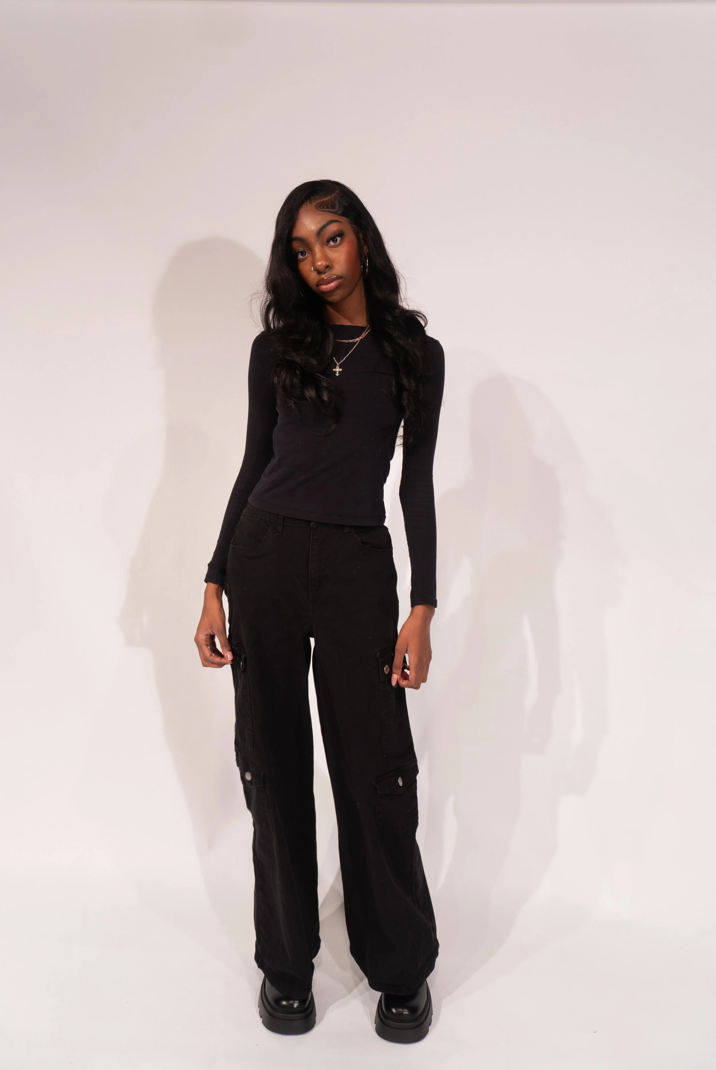 A young woman with dark hair styled in loose waves, wearing a black long-sleeve top, black cargo pants, and black platform shoes, standing against a plain white background.
