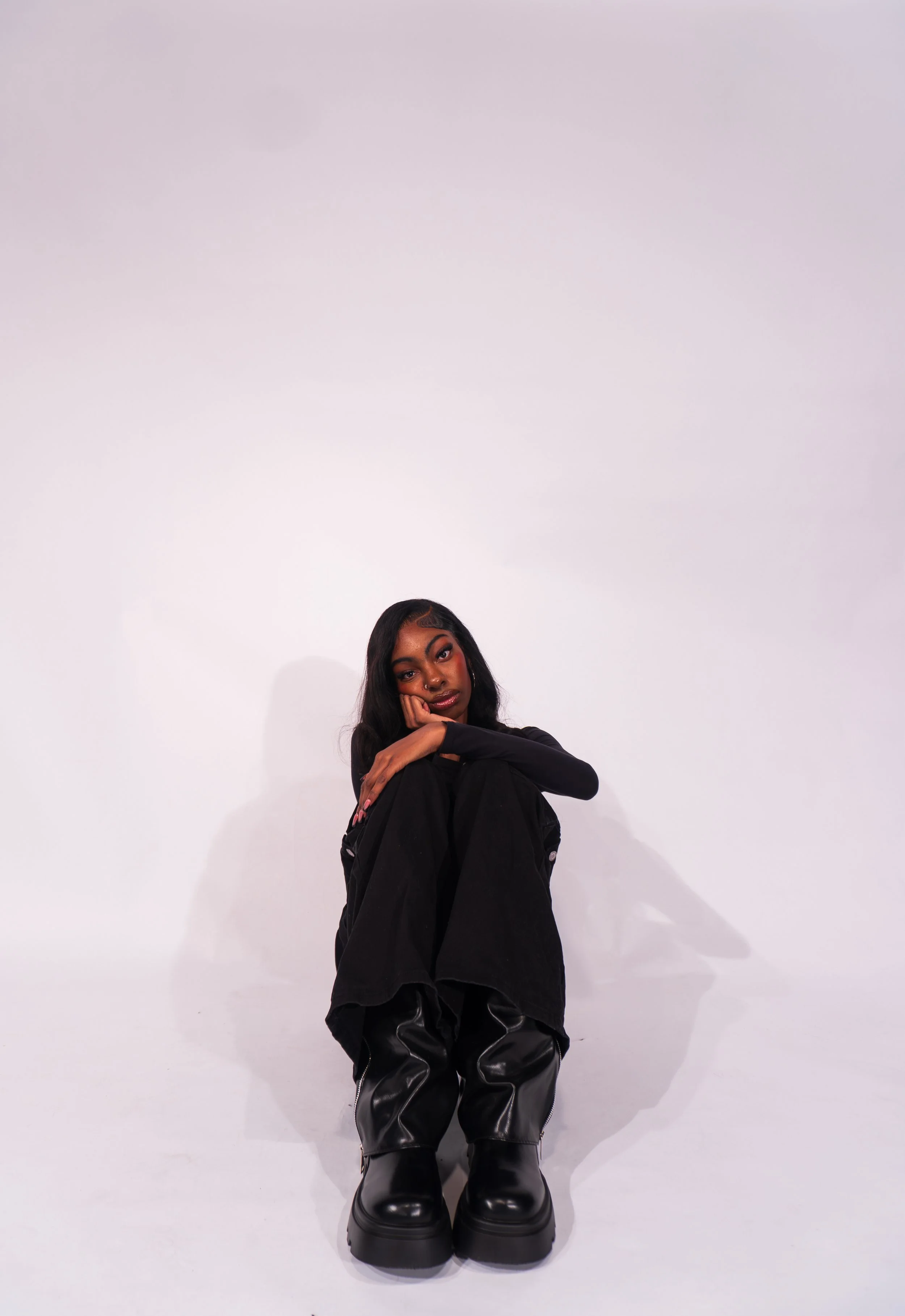 Young woman sitting on the floor, resting her chin on her knees, wearing black clothing and chunky black boots, against a plain white background.