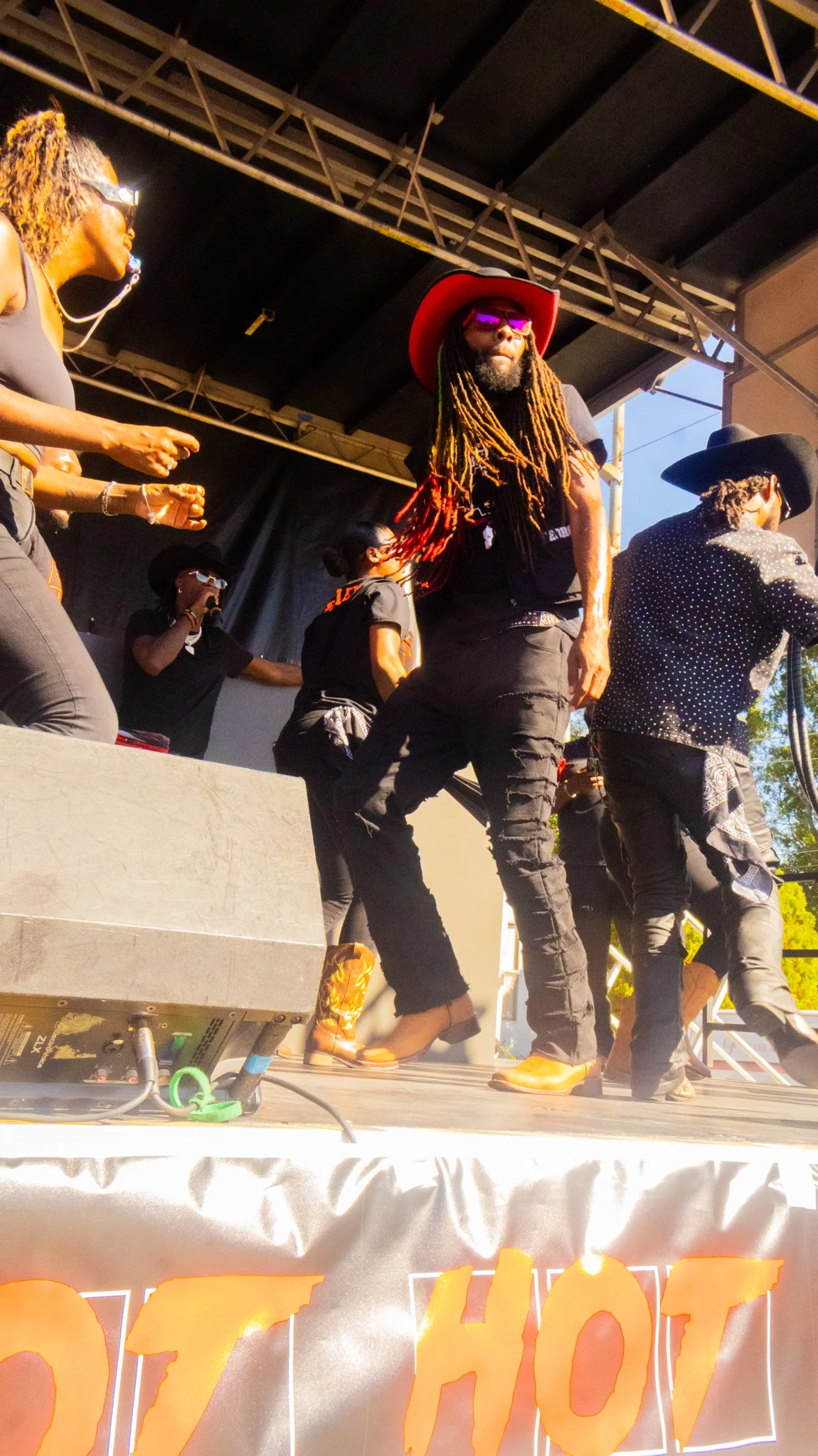 A group of musicians performing on stage during a live outdoor concert, with one man in a red hat, sunglasses, and dreadlocks dancing in front.
