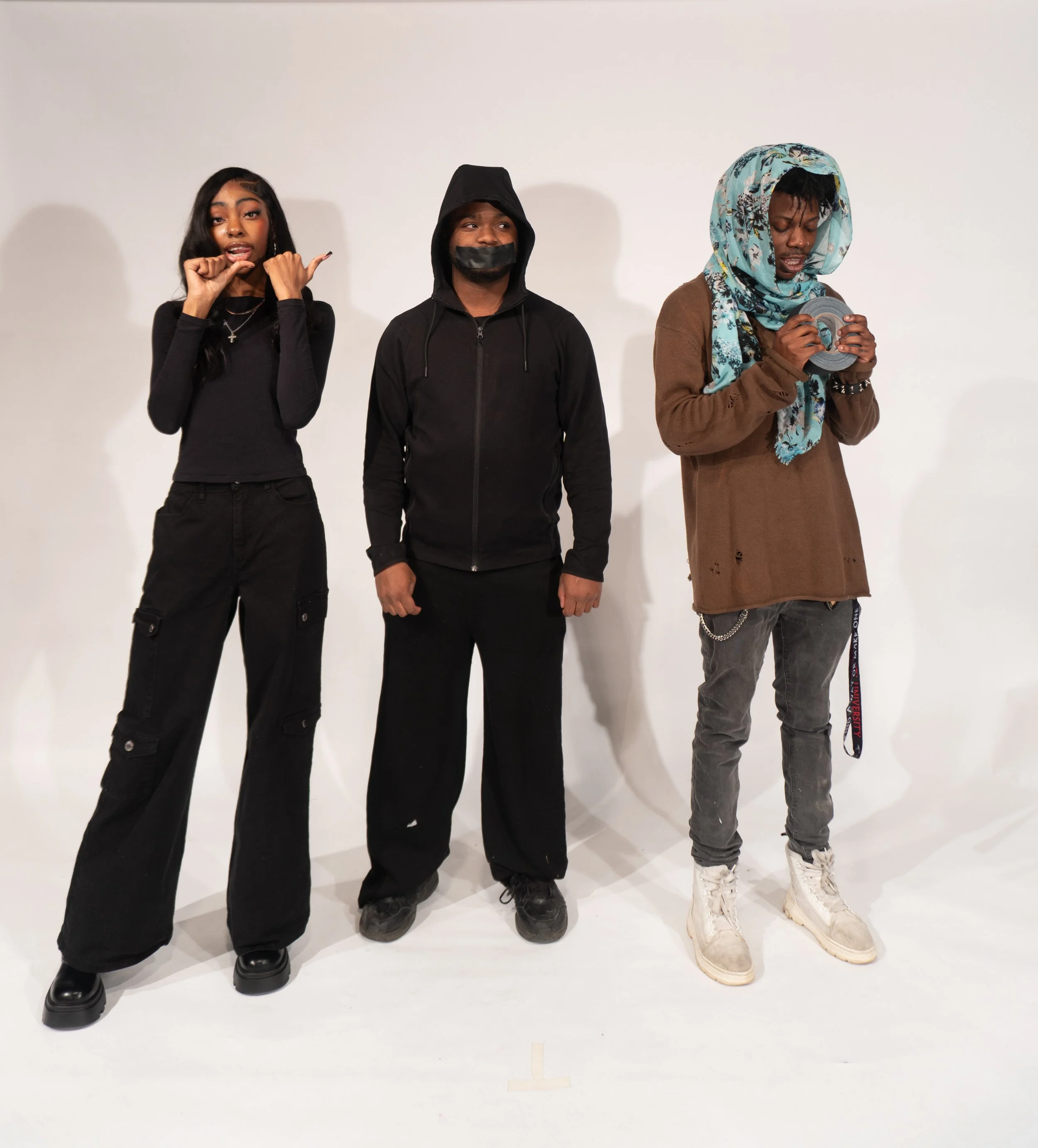 Three young people standing side by side against a plain white background, one woman and two men, dressed in casual streetwear, with the woman holding her hand near her face, the middle man wearing a hoodie and face mask, and the man on the right wearing a headscarf and holding a roll of tape.