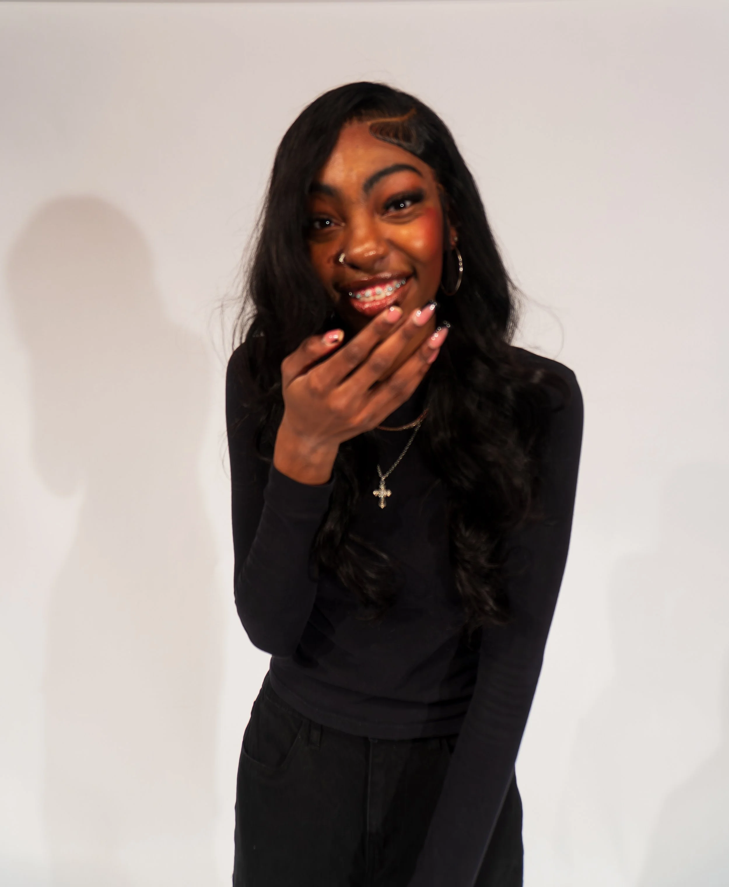 A young woman with long black hair, wearing a black long-sleeve shirt, smiling and covering her mouth with her hand, standing against a plain white background.