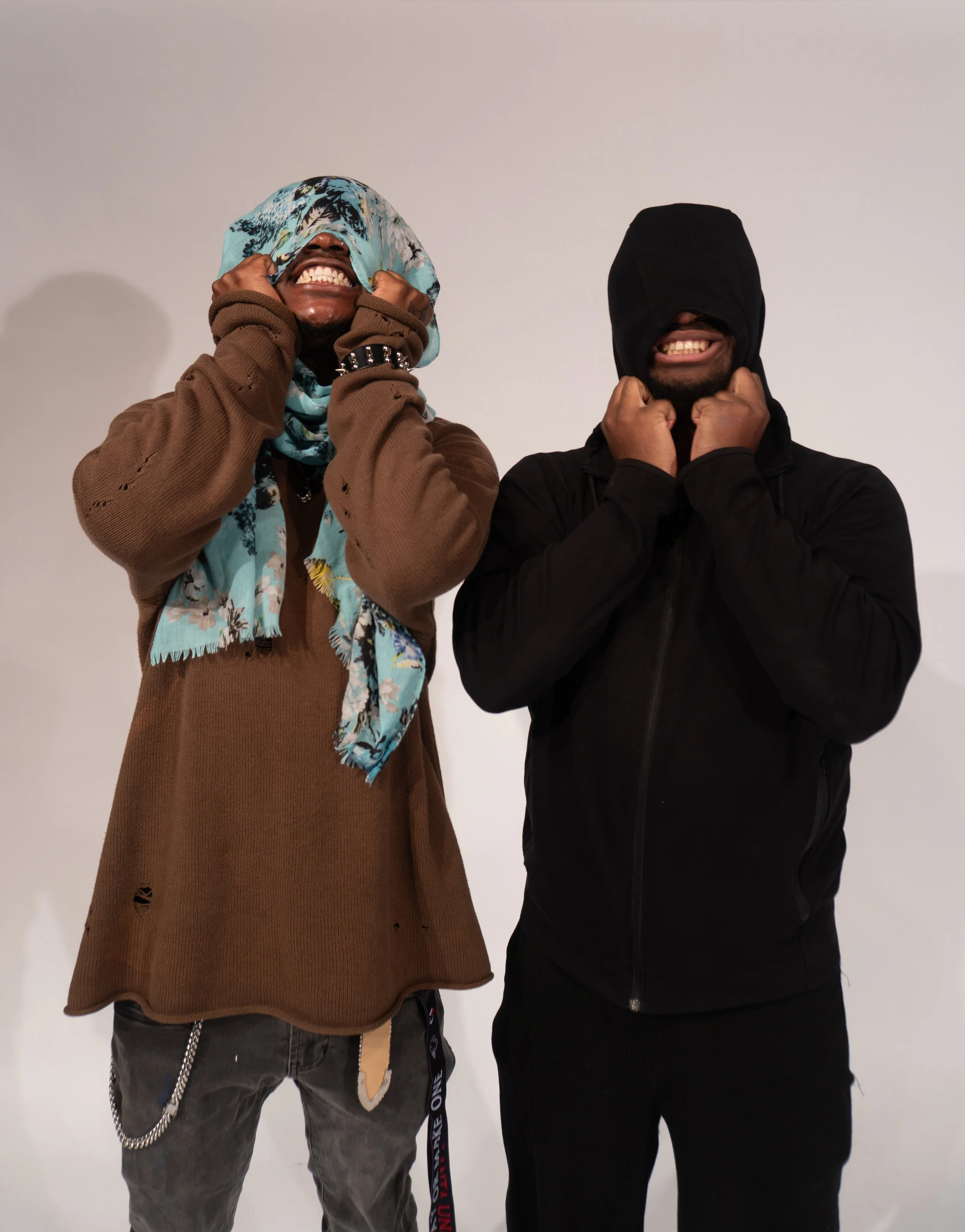 Two men stand side by side, both covering their heads with their hoodies and showing their teeth in playful expressions against a plain white background.