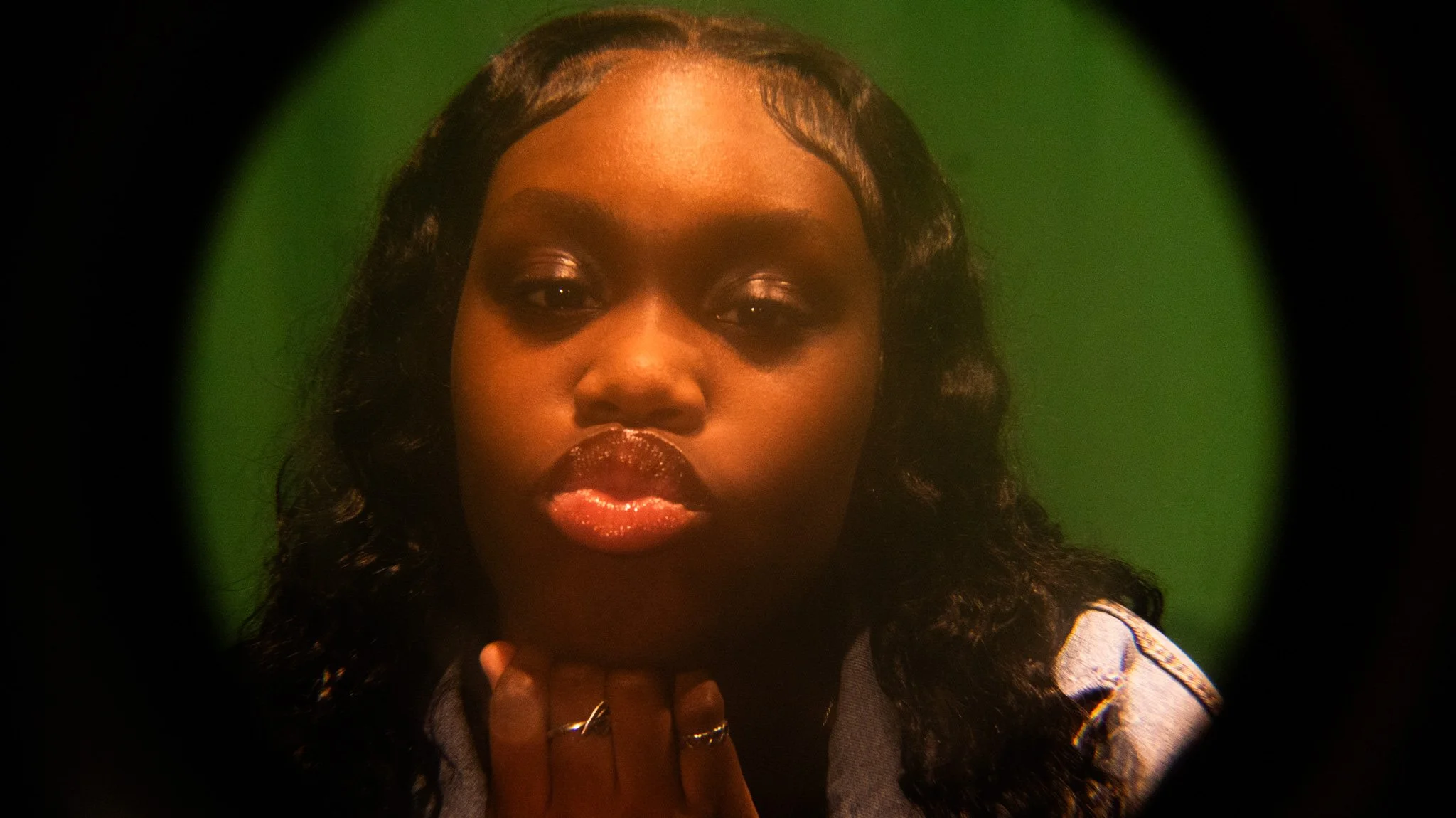 Close-up of a young woman with dark curly hair, glossy lips, and wearing rings, against a green background, viewed through a circular lens.