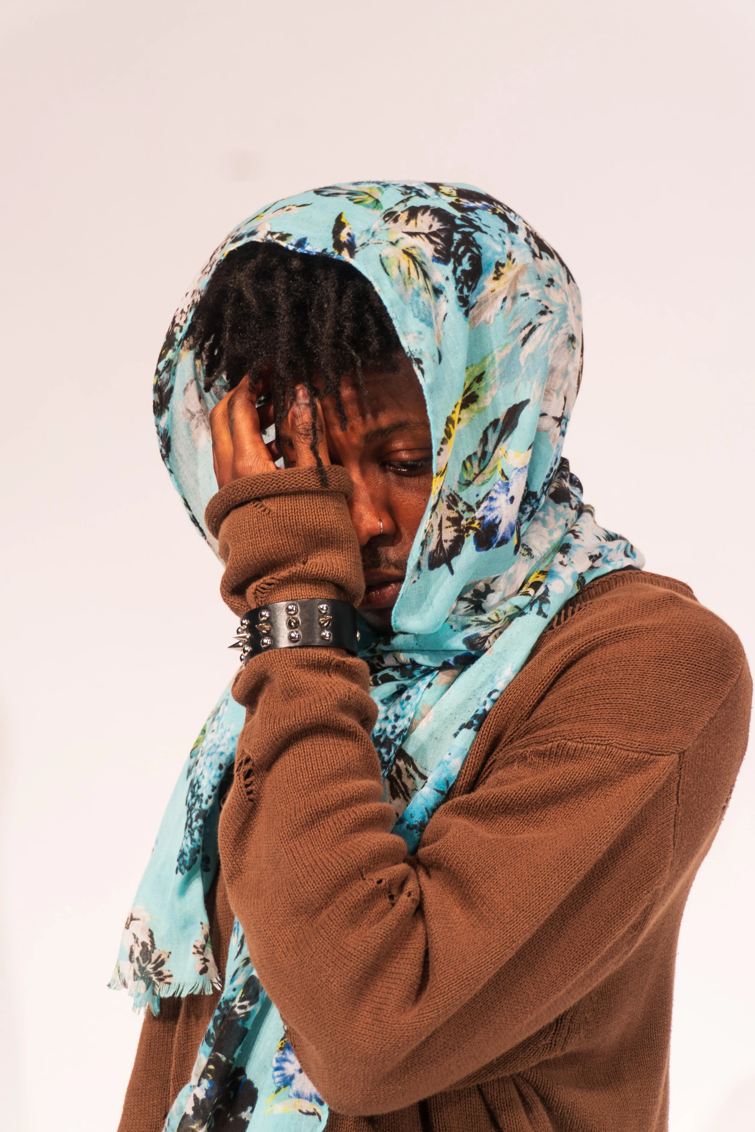 A person with dreadlocks, wearing a floral headscarf and a brown sweater, holding one side of their head with a pained expression.