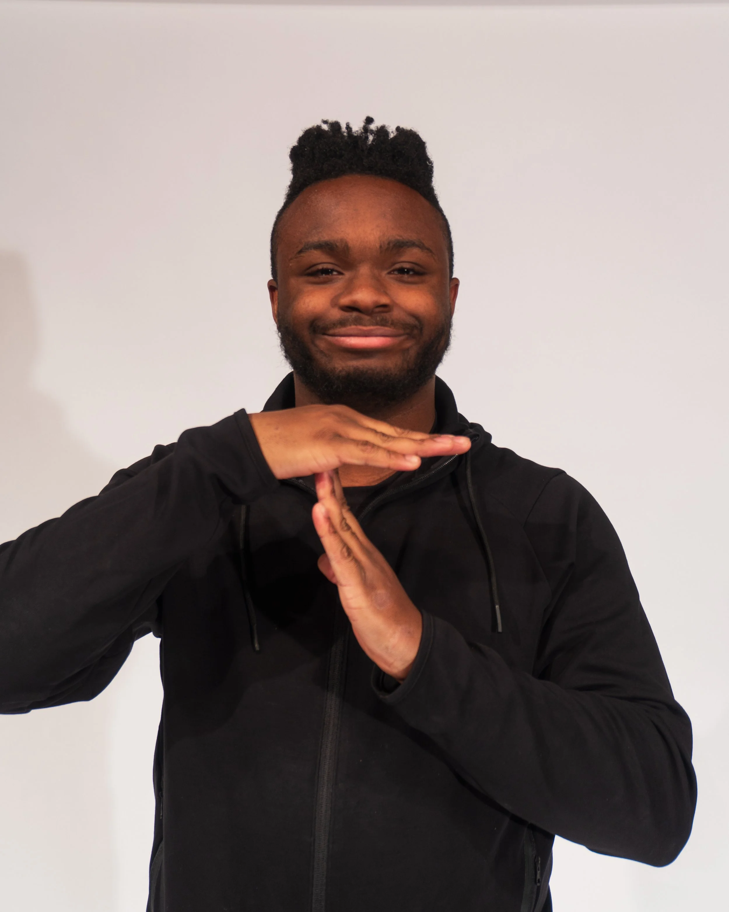 A young man with short curly hair and a beard smiling, wearing a black hoodie, making a gesture with his hands against a plain white background.