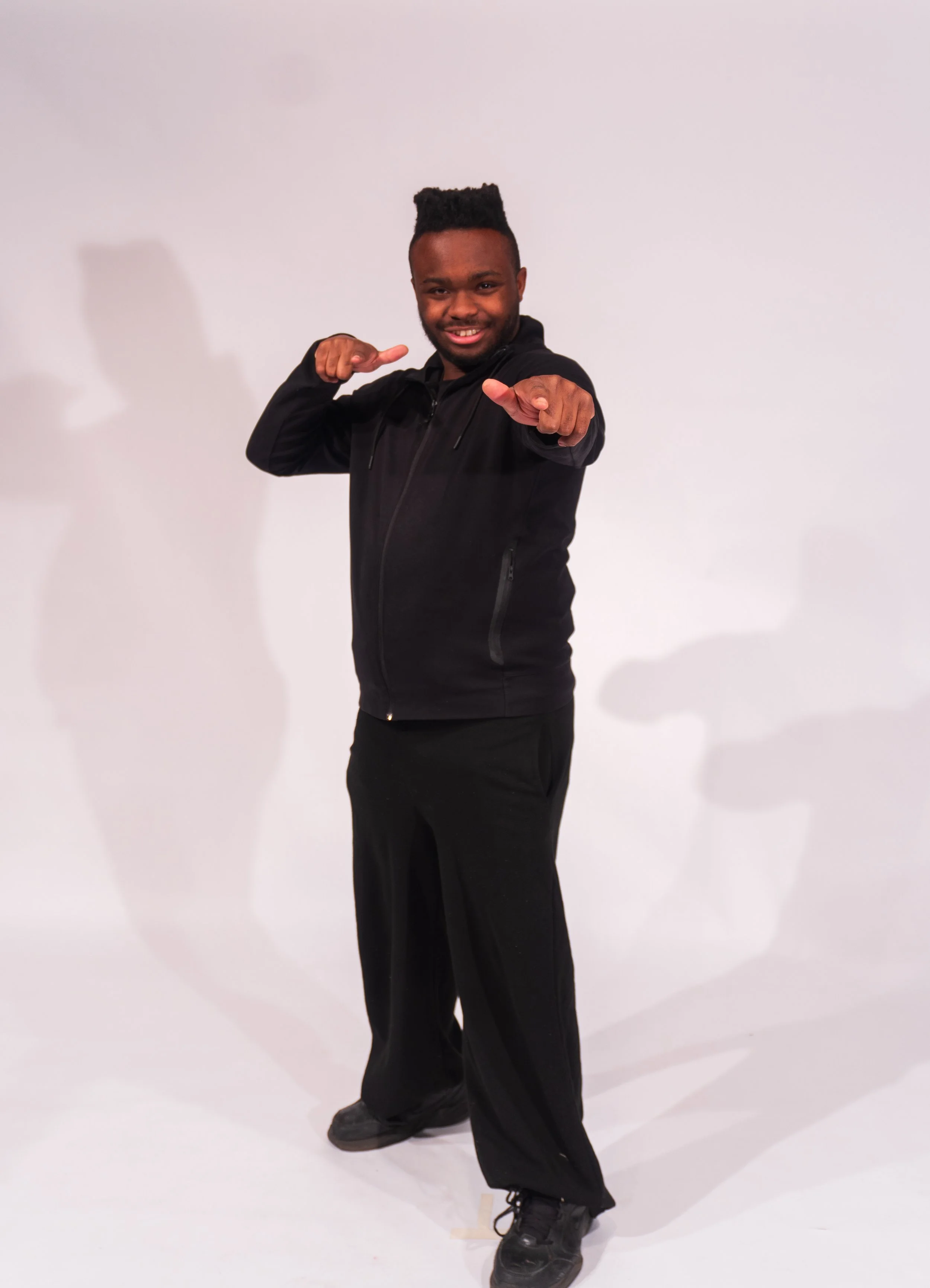 A man with dark skin, a tall flat-top hairstyle, wearing a black jacket, black pants, and black shoes, standing against a white background, smiling and pointing towards the camera.