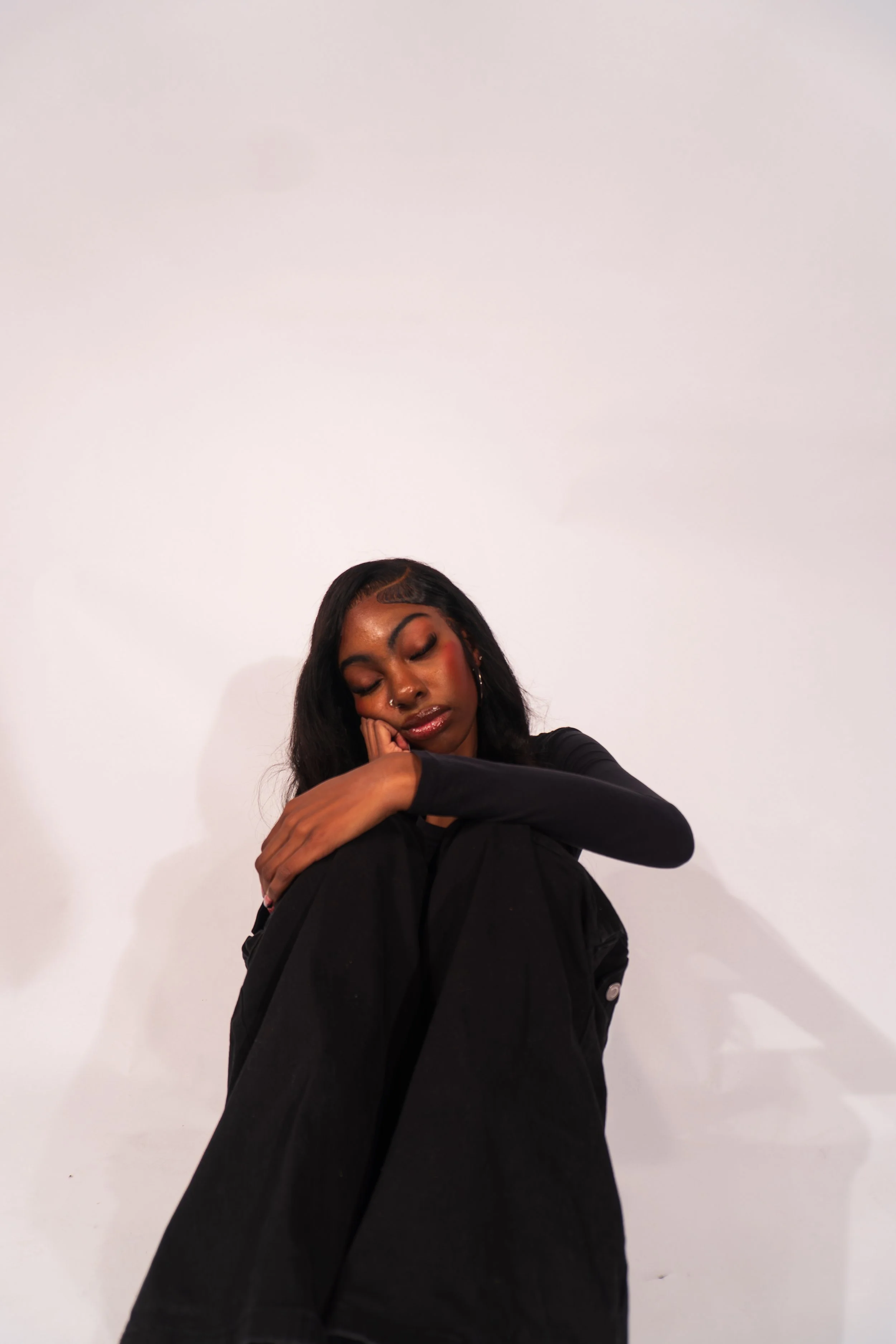 Young woman with dark hair and makeup sitting with her head resting on her knees, eyes closed, against a plain white background.