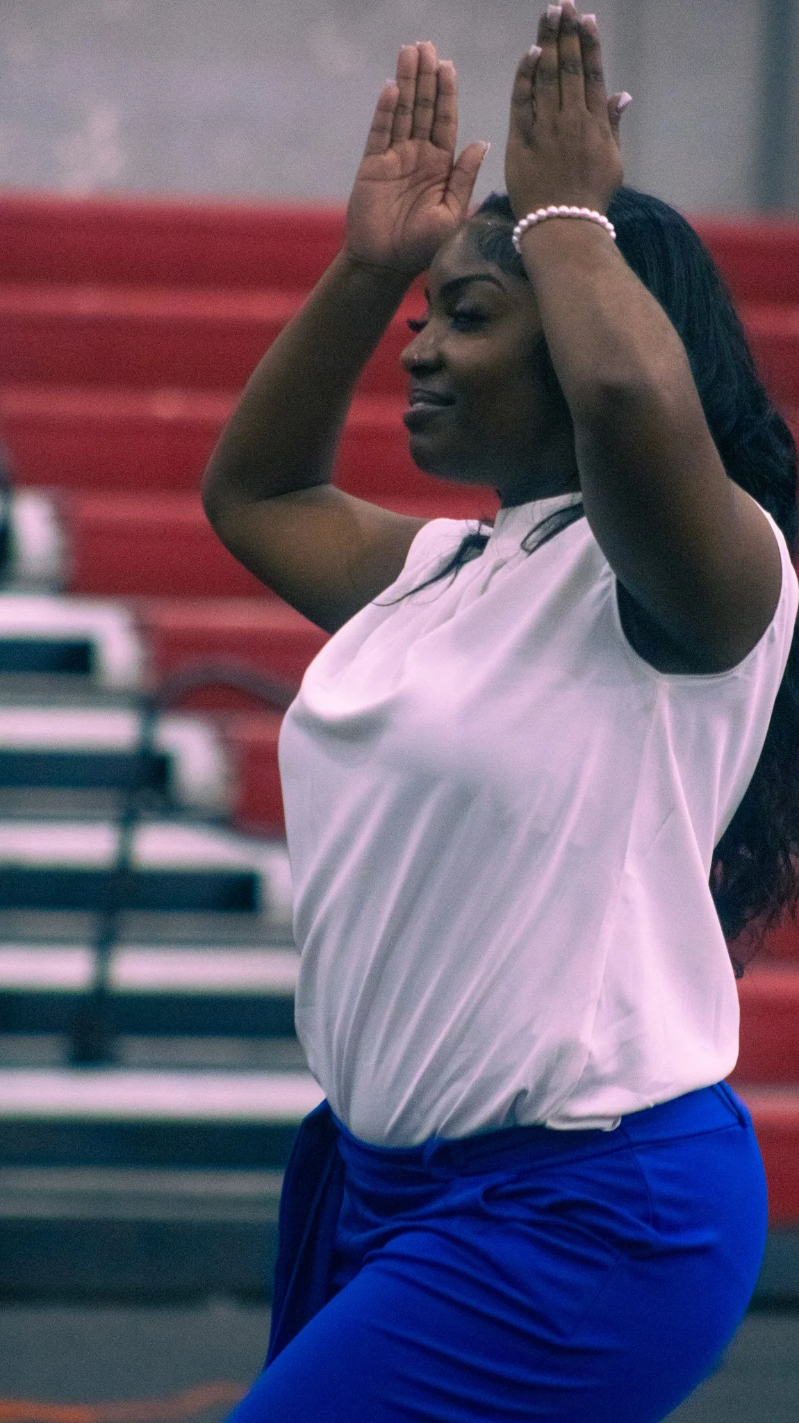A woman with dark hair wearing a white shirt, blue pants, a pink bracelet, and pink and white nails, standing with her hands raised above her head in a gym or sports facility.