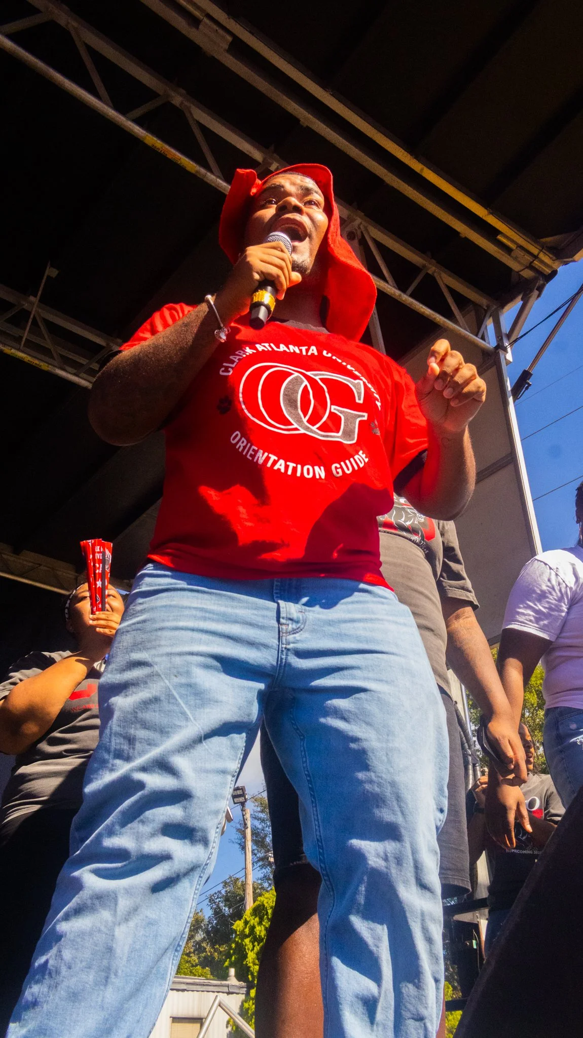 A person speaking at a rally or event, wearing a red T-shirt and a red hat, holding a microphone, with other people in the background, and a stage structure above.