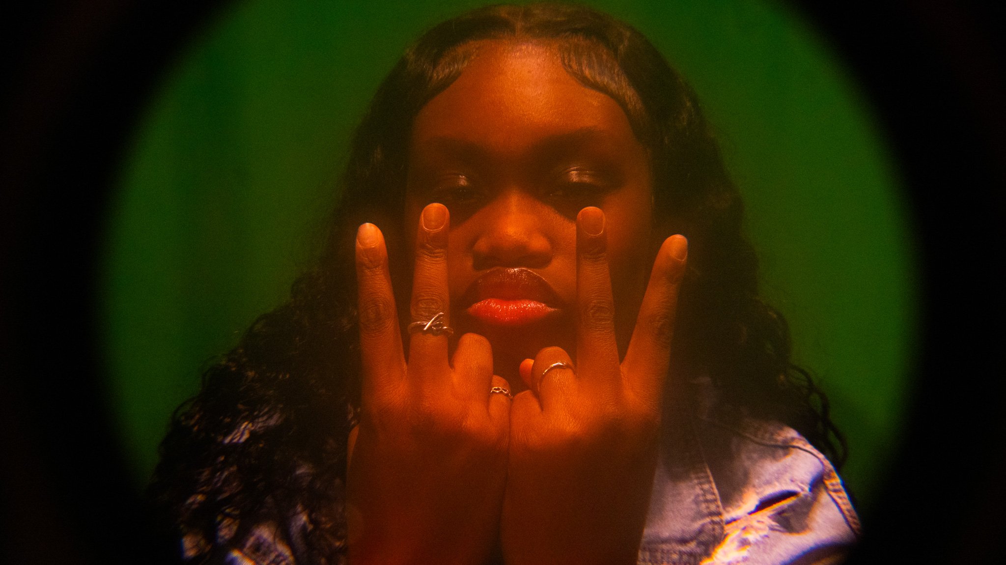 A woman with curly dark hair holding up her hand with fingers forming a peace sign in front of her face, against a green background.