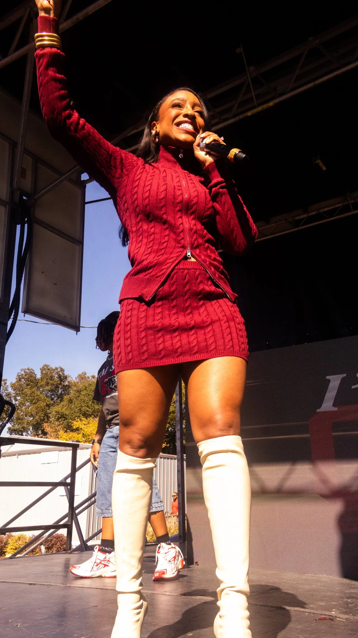 A woman in a red cable-knit sweater dress and white thigh-high boots holds a microphone, smiling confidently on stage during an outdoor event.