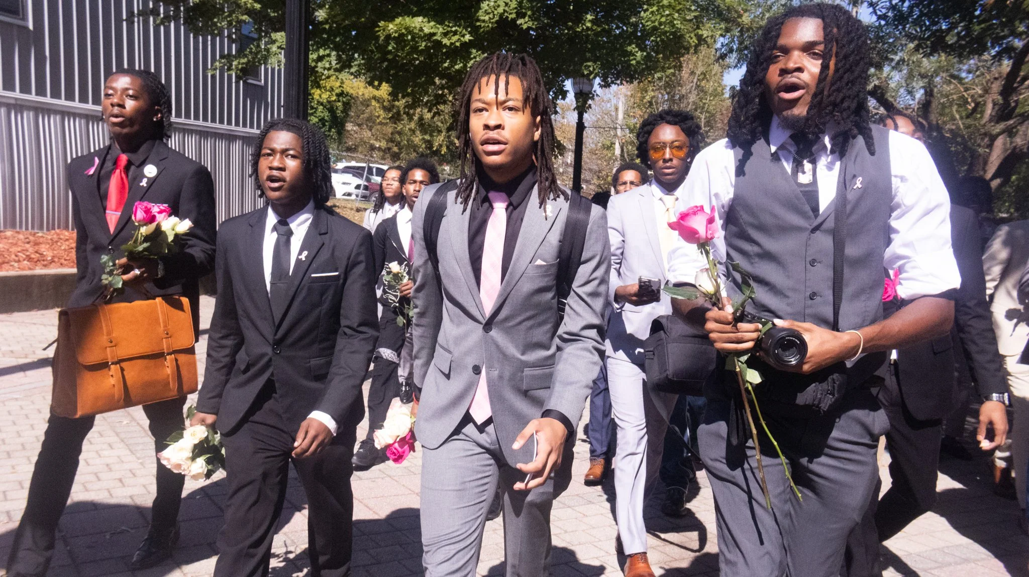 Group of people participating in a march or demonstration, some holding pink roses and wearing formal attire with ribbons, outdoors on a sunny day.
