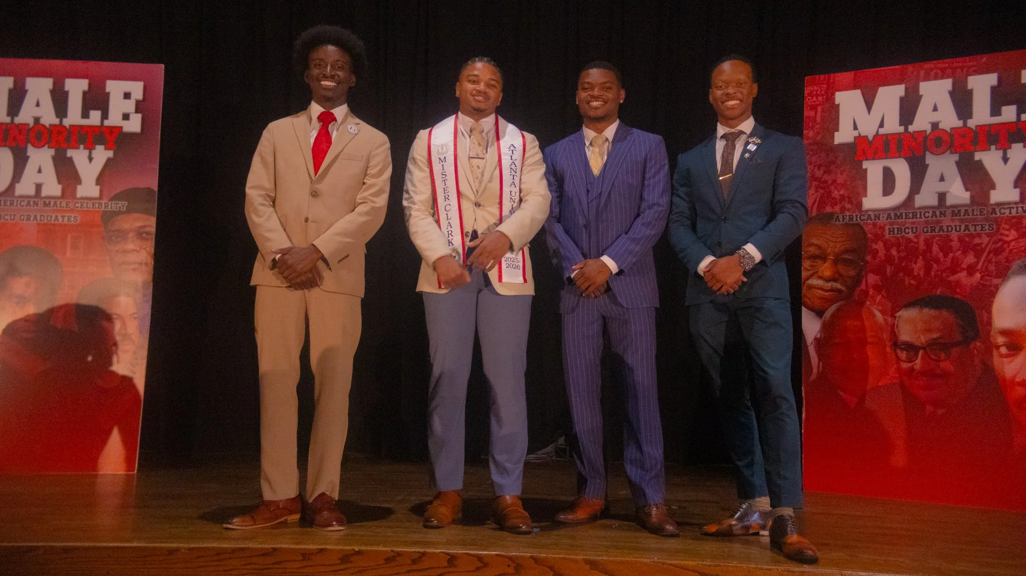 Five young men dressed in formal suits standing on stage at an event, with two red banners on each side. The event celebrates African-American male active graduates. The second person from the left is wearing a sash that reads "MISTER CLARK" and a ribbon with the years 2025-2026.