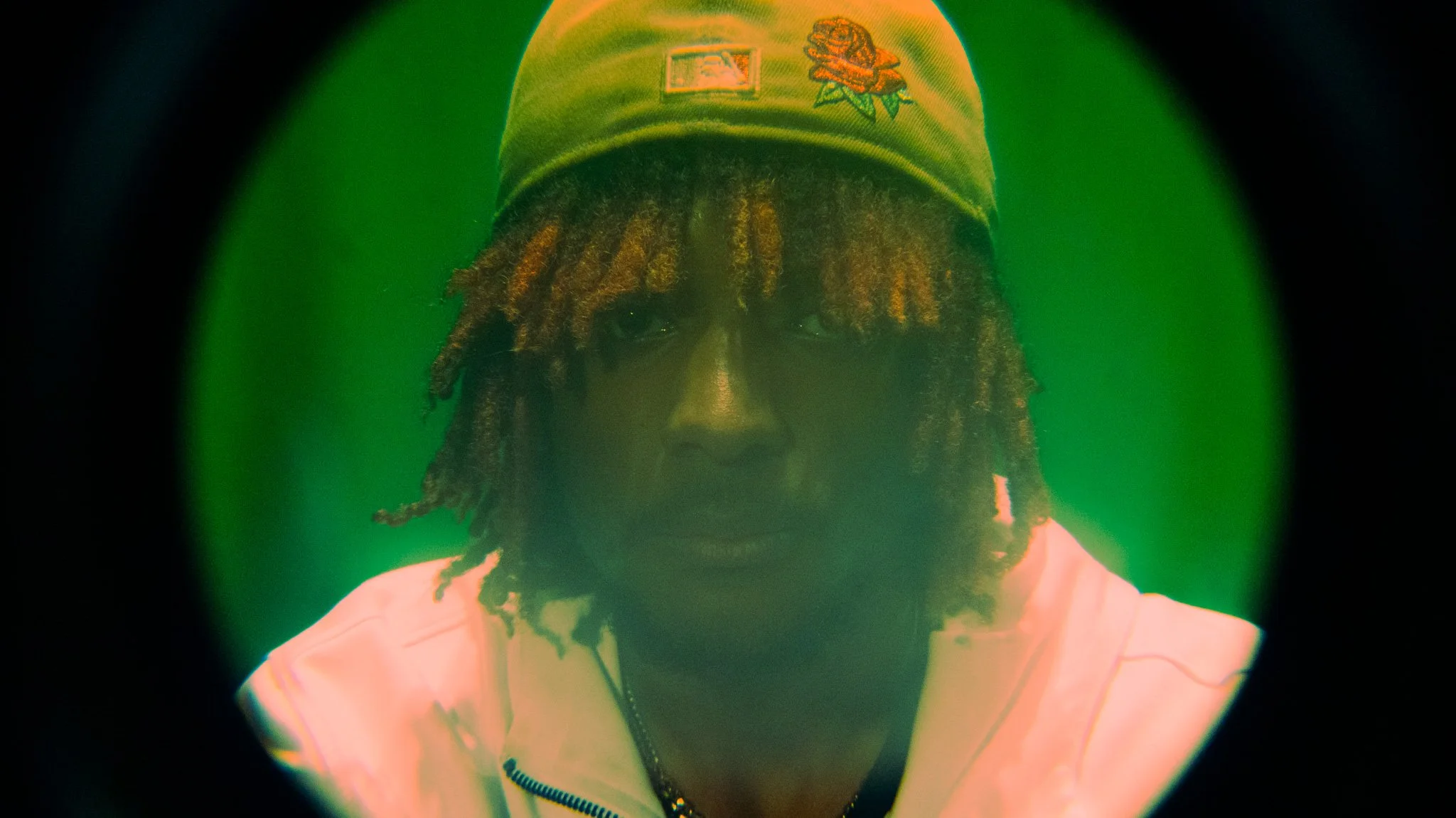 A person with dreadlocks wearing a beige cap with a rose embroidery on it, looking directly at the camera. The photo has a green background and is taken through a circular frame.