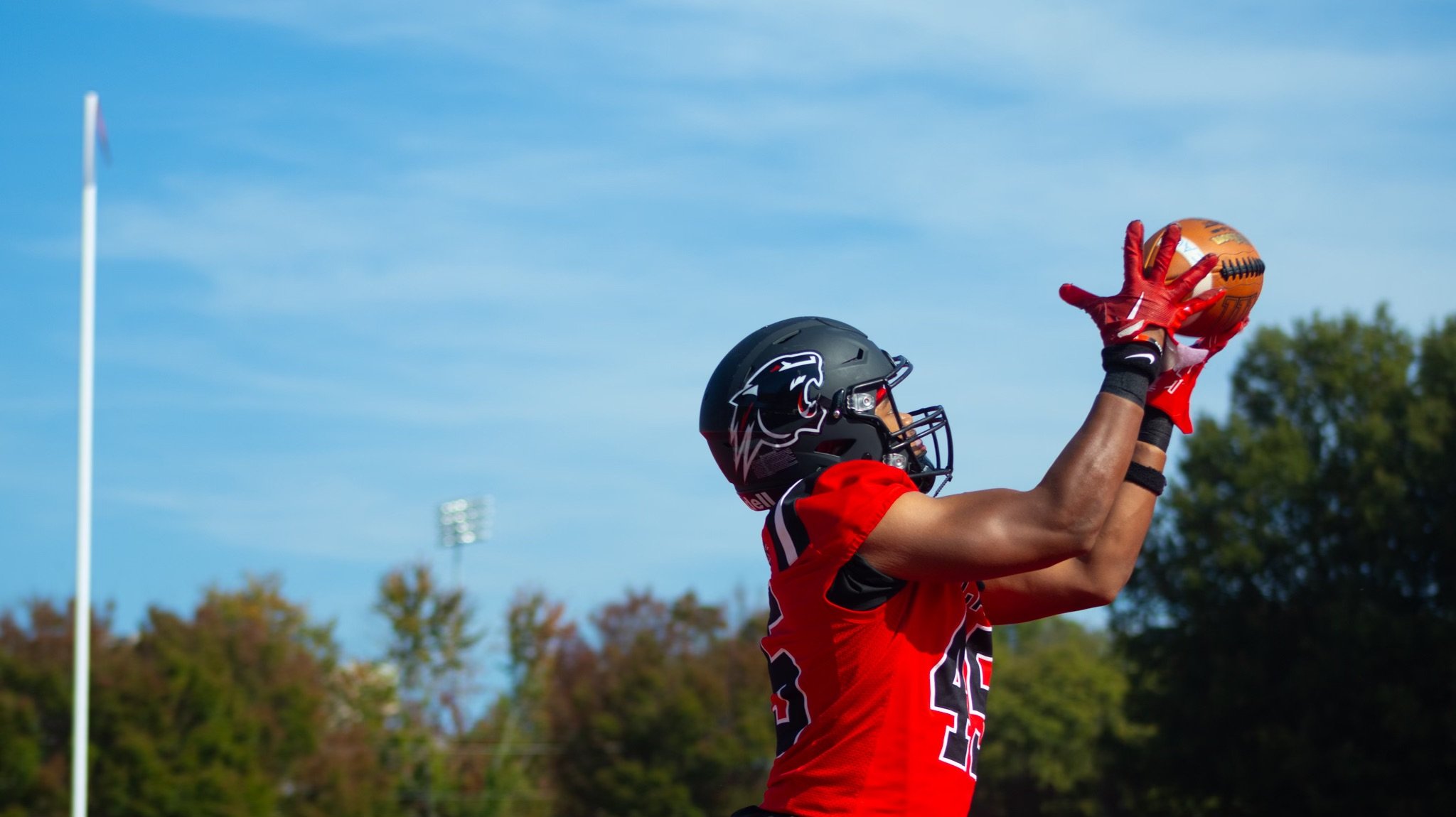 Football player in red jersey and black helmet catching a football during a game outdoors on a clear day.
