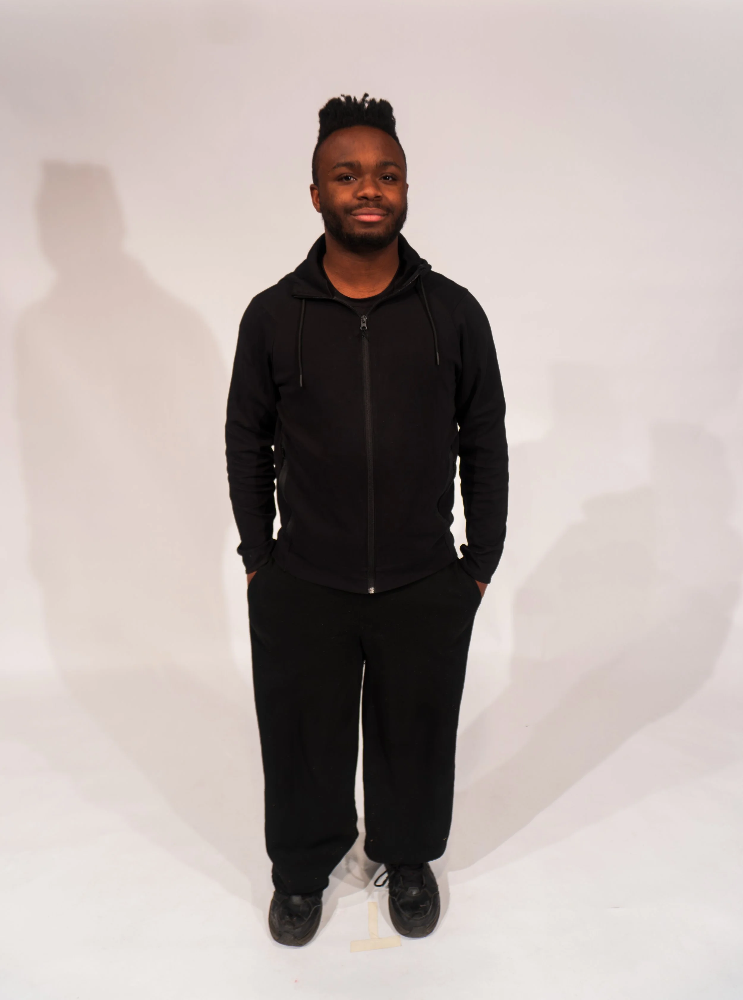 A young man with dark skin and a hairstyle with short sides and a taller top, wearing a black zip-up hoodie, black pants, and black sneakers, standing against a plain white background with his hands in his pockets.