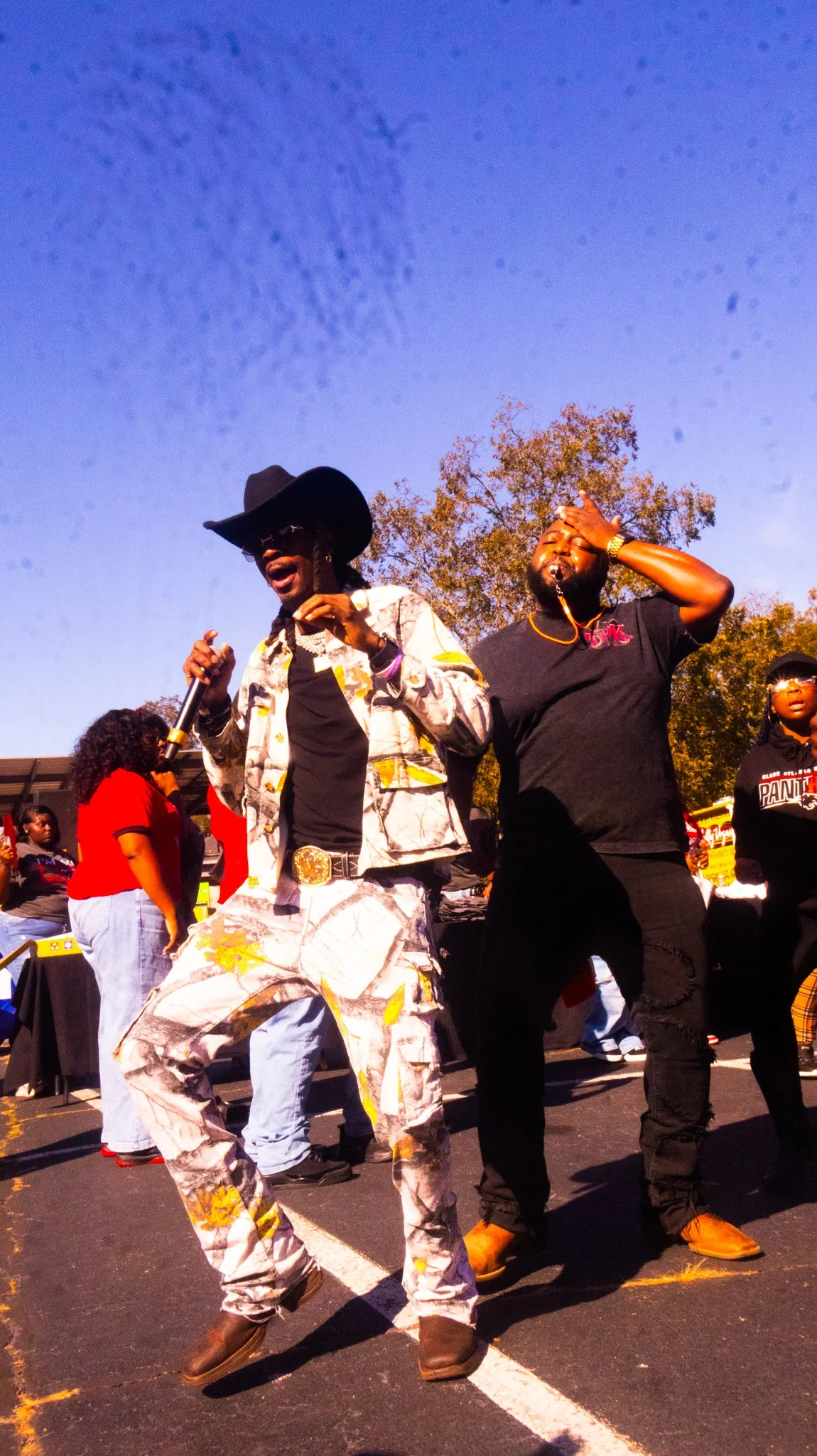 Two men dancing outdoors at a gathering, one wearing a cowboy hat and patterned outfit singing with a microphone, the other wearing a black shirt with a logo, in front of a crowd.