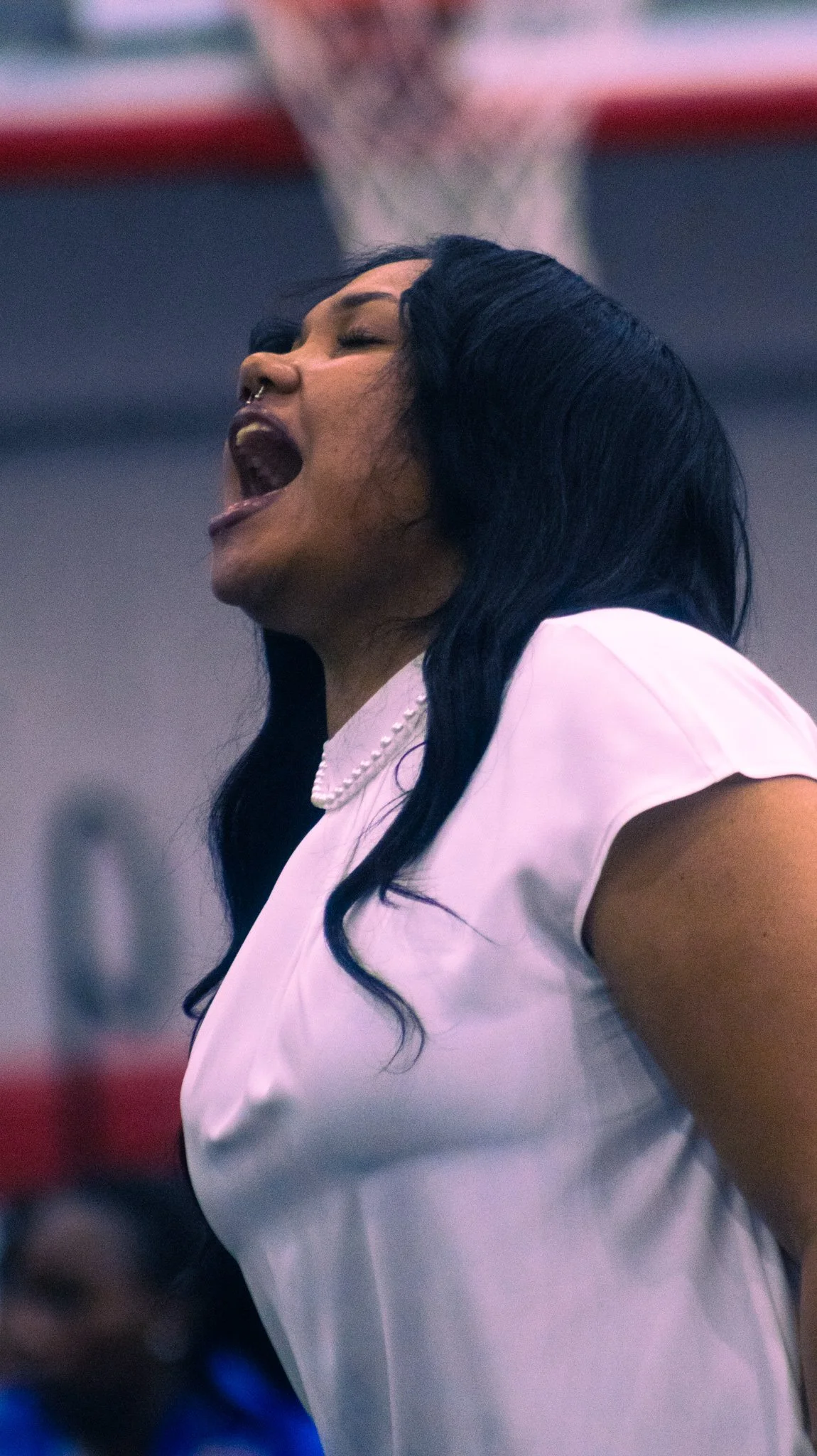 A woman with long dark hair, wearing a white top and a pearl necklace, appears to be singing or shouting with her mouth wide open.