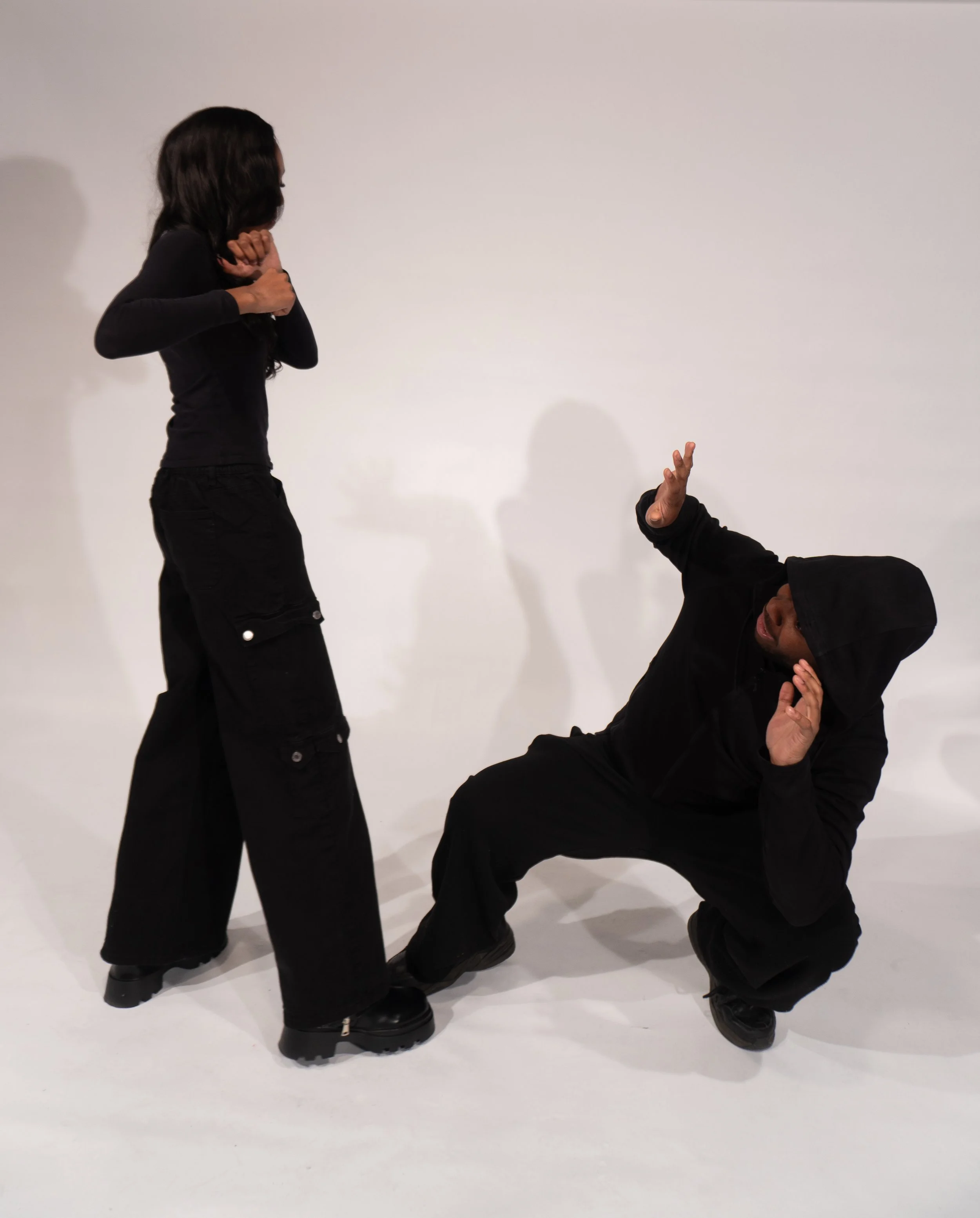 Two individuals dressed in black, one standing with hands near face and the other kneeling on one knee with one arm raised, appear to be performing a dramatic scene against a plain white background.