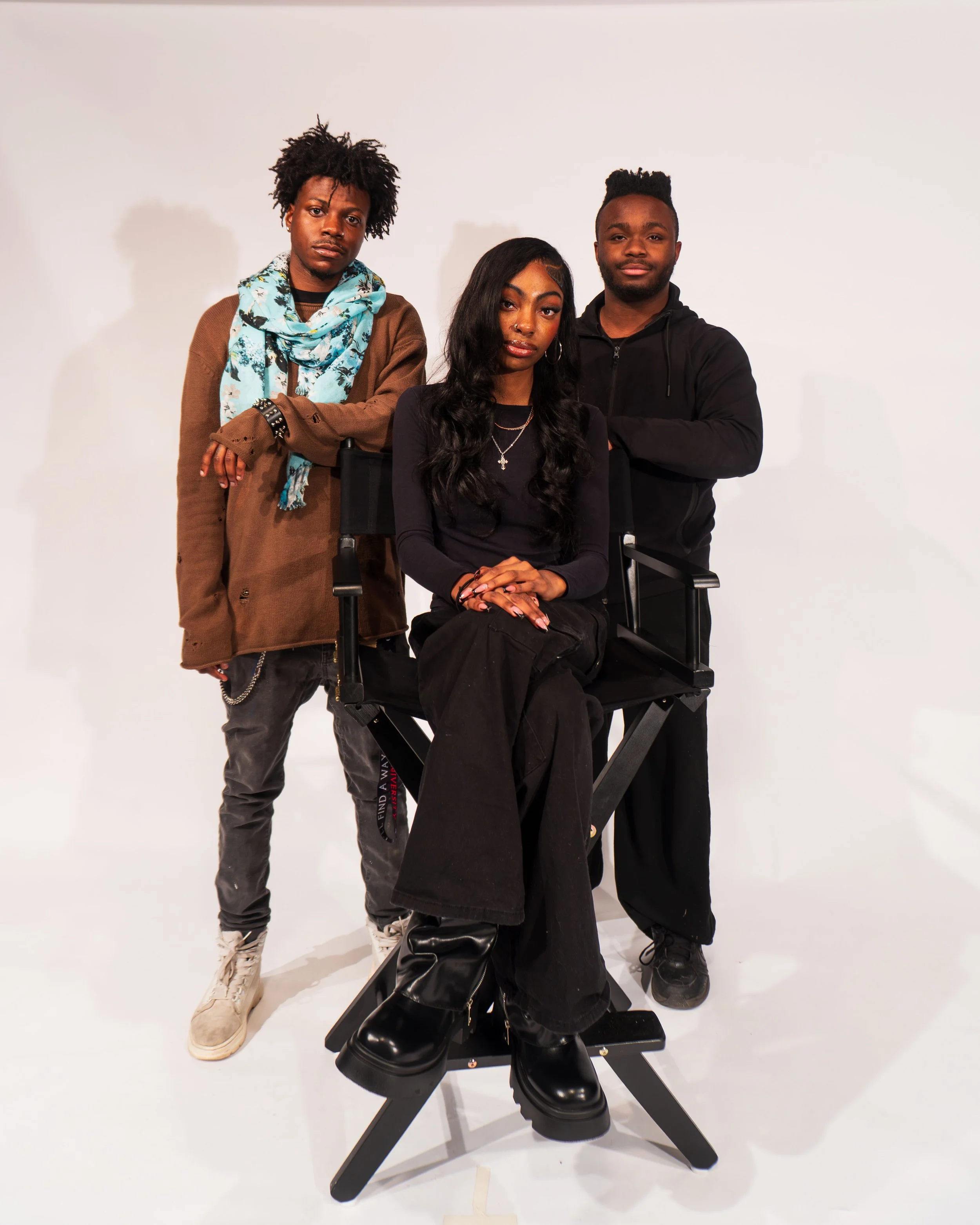 Three Black individuals, two men and one woman, posing against a white background. The woman is seated on a director's chair, wearing black clothes and chunky black boots, with long wavy hair. The man to her left has curly hair, casual brown jacket, and scarf, and the man to her right has short hair, beard, and arm crossed, wearing black hoodie.