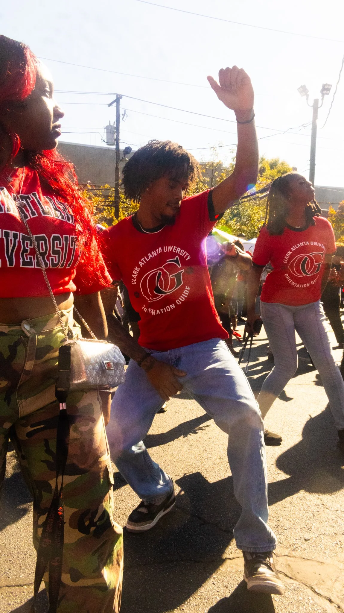 A group of young women dancing outdoors on a sunny day, wearing red shirts with university and orientation guide logos, surrounded by other people.