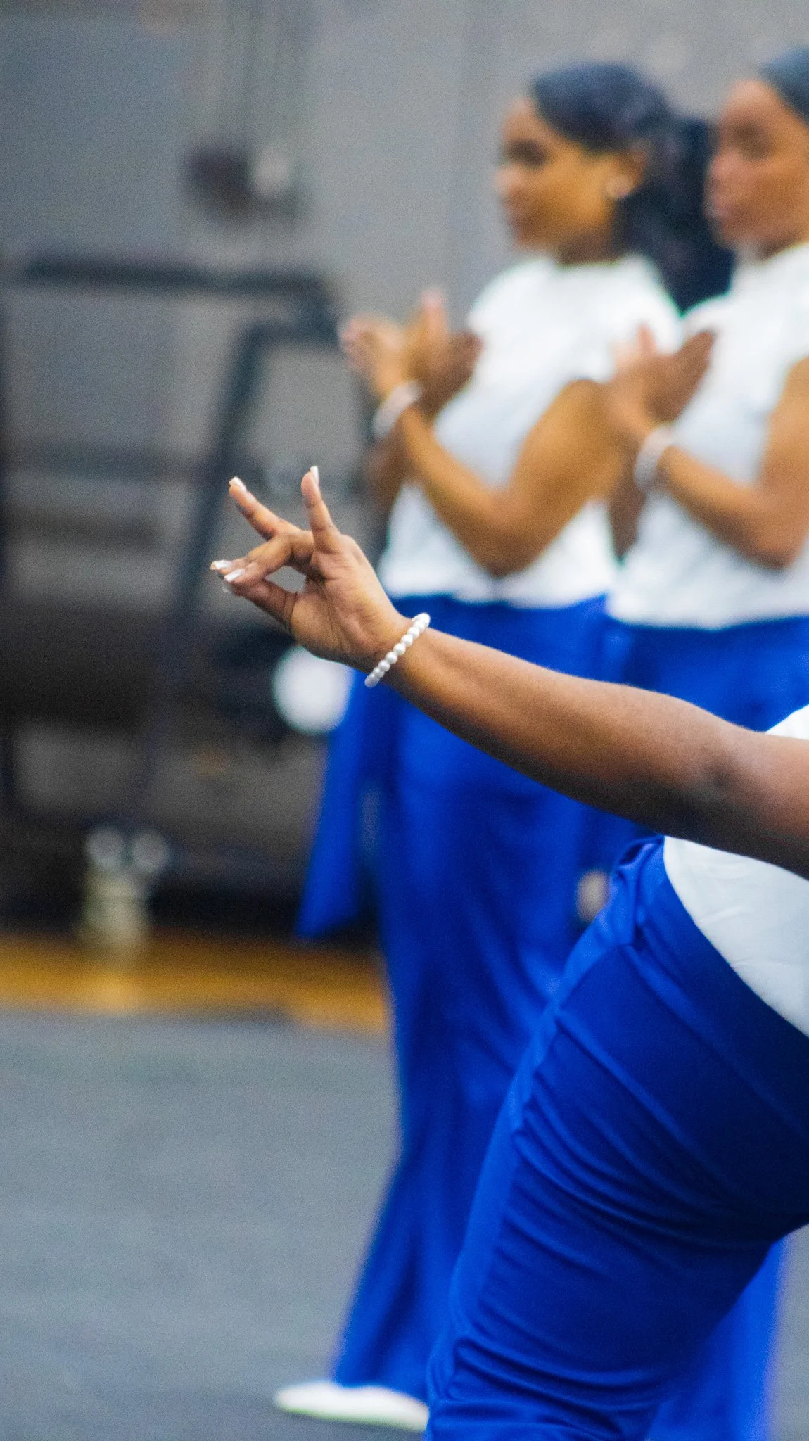 Close-up of a woman in blue pants and a white top practicing yoga, with her hand forming a mudra, in a gym or studio setting.