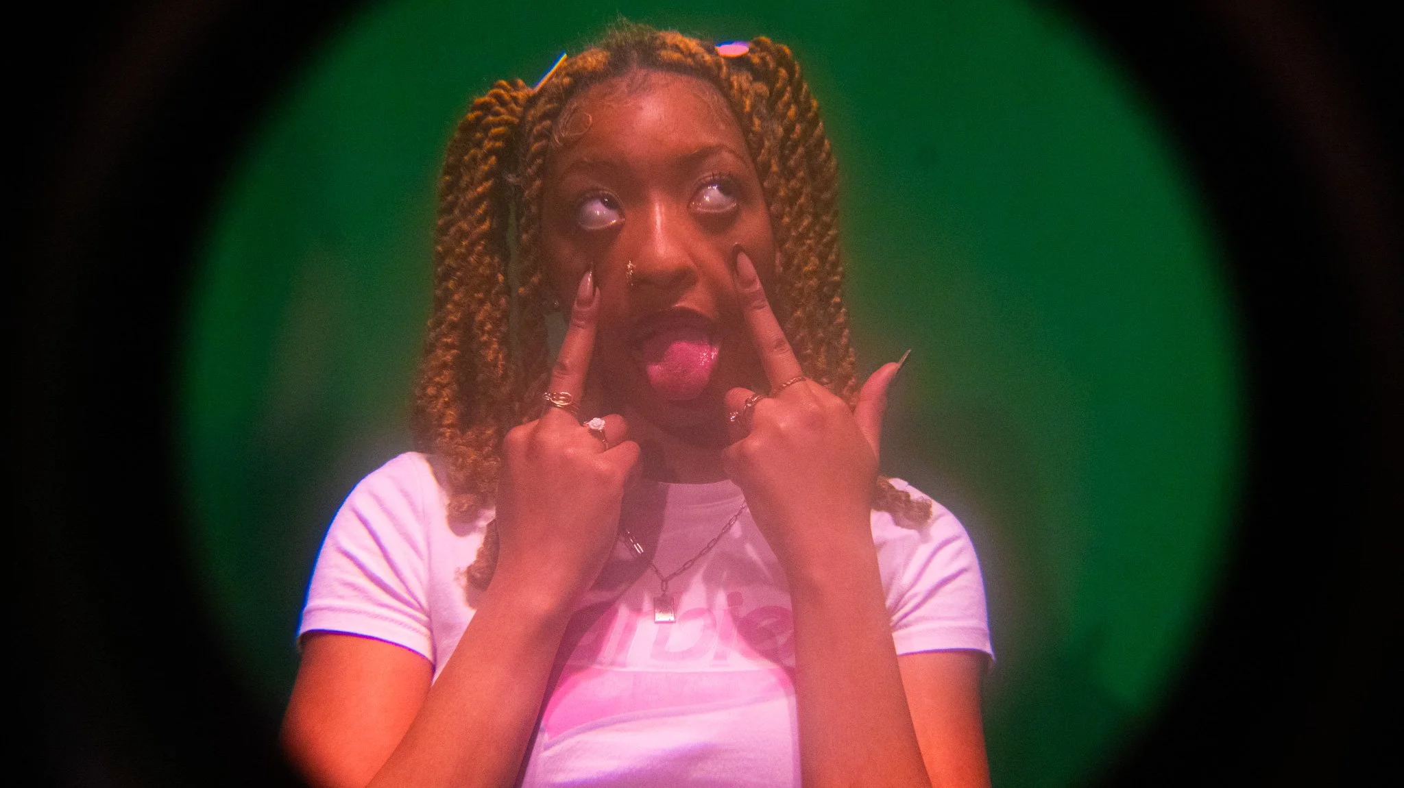 Young woman with braided hair making playful face, sticking out her tongue and holding her face with both hands under green lighting