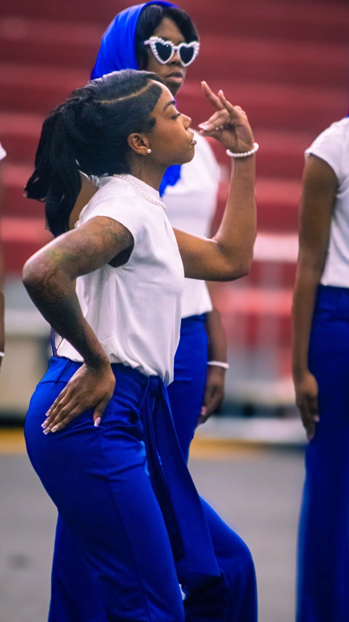 Two women are shown mid-dance in a gymnasium or sports hall, with one woman in the foreground wearing a white top, blue sweatpants, and pearl accessories, raising her hand with fingers positioned to suggest a dance move. The woman in the background is wearing heart-shaped sunglasses, a blue headscarf, and white top, observing or participating in the dance.