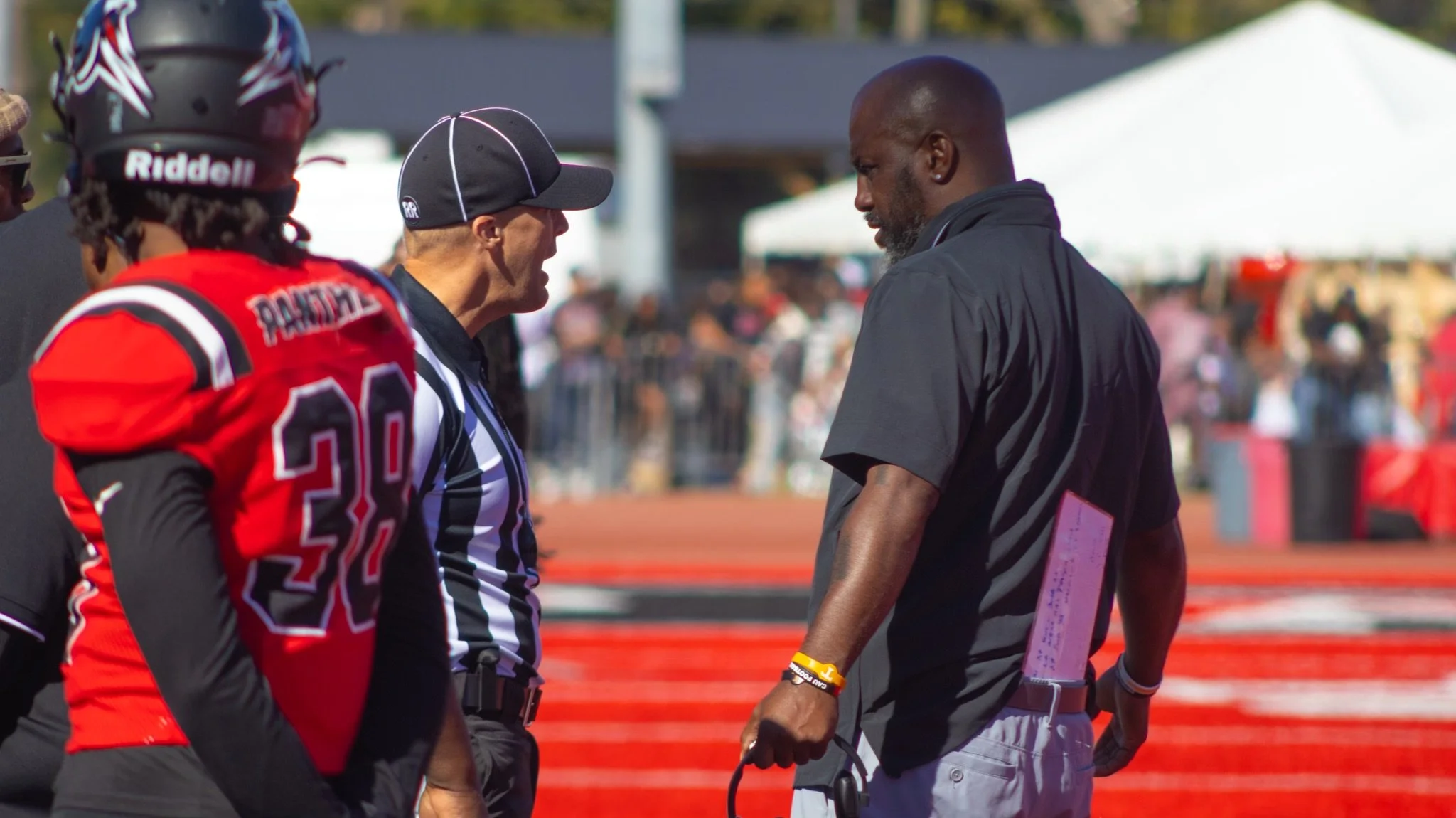 Two football players and two officials having a conversation on the field during a game or event, with a crowd and tents in the background.