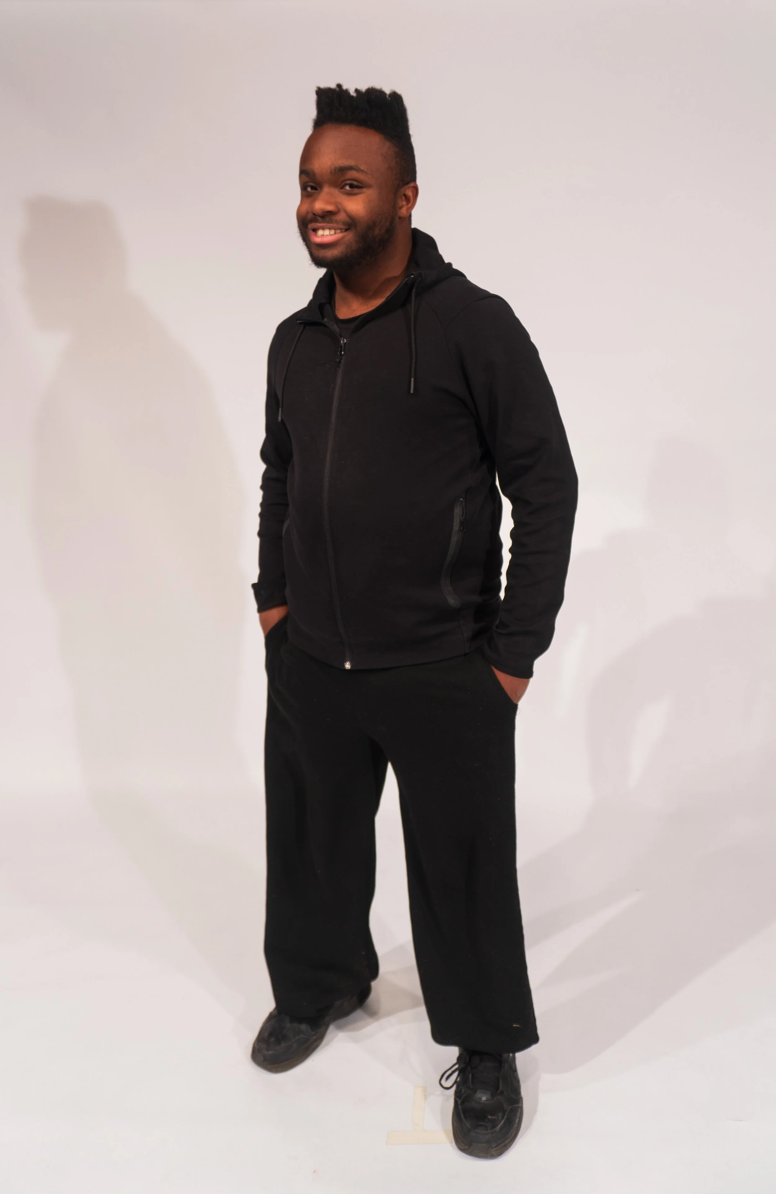 A young man with dark skin, a beard, and a high-top hairstyle, wearing a black zip-up hoodie, black pants, and black shoes, standing with hands in pockets against a plain white background.