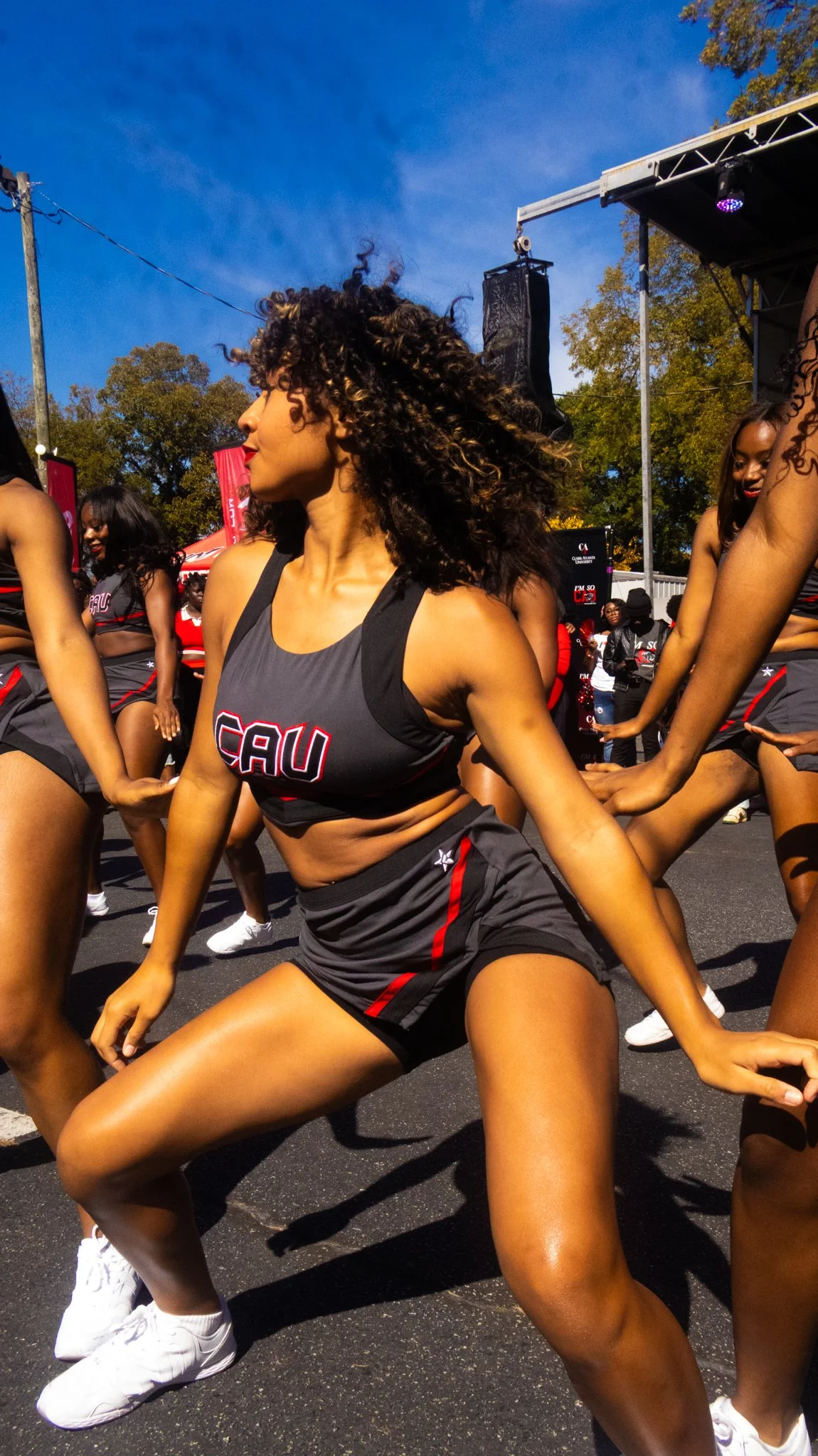 Cheerleaders performing a dance routine outdoors during a parade or event, wearing black and red uniforms with 'CAU' written on them, under a clear blue sky.
