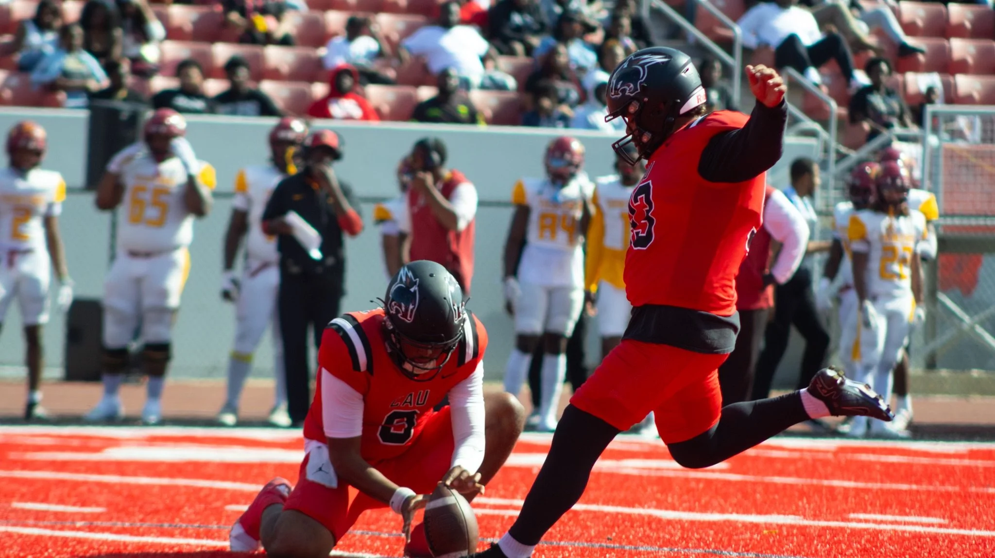 A football player in a red uniform kneeling and holding a football, with another player in a red and black uniform running near him on the field. Spectators are watching from the stands in the background.
