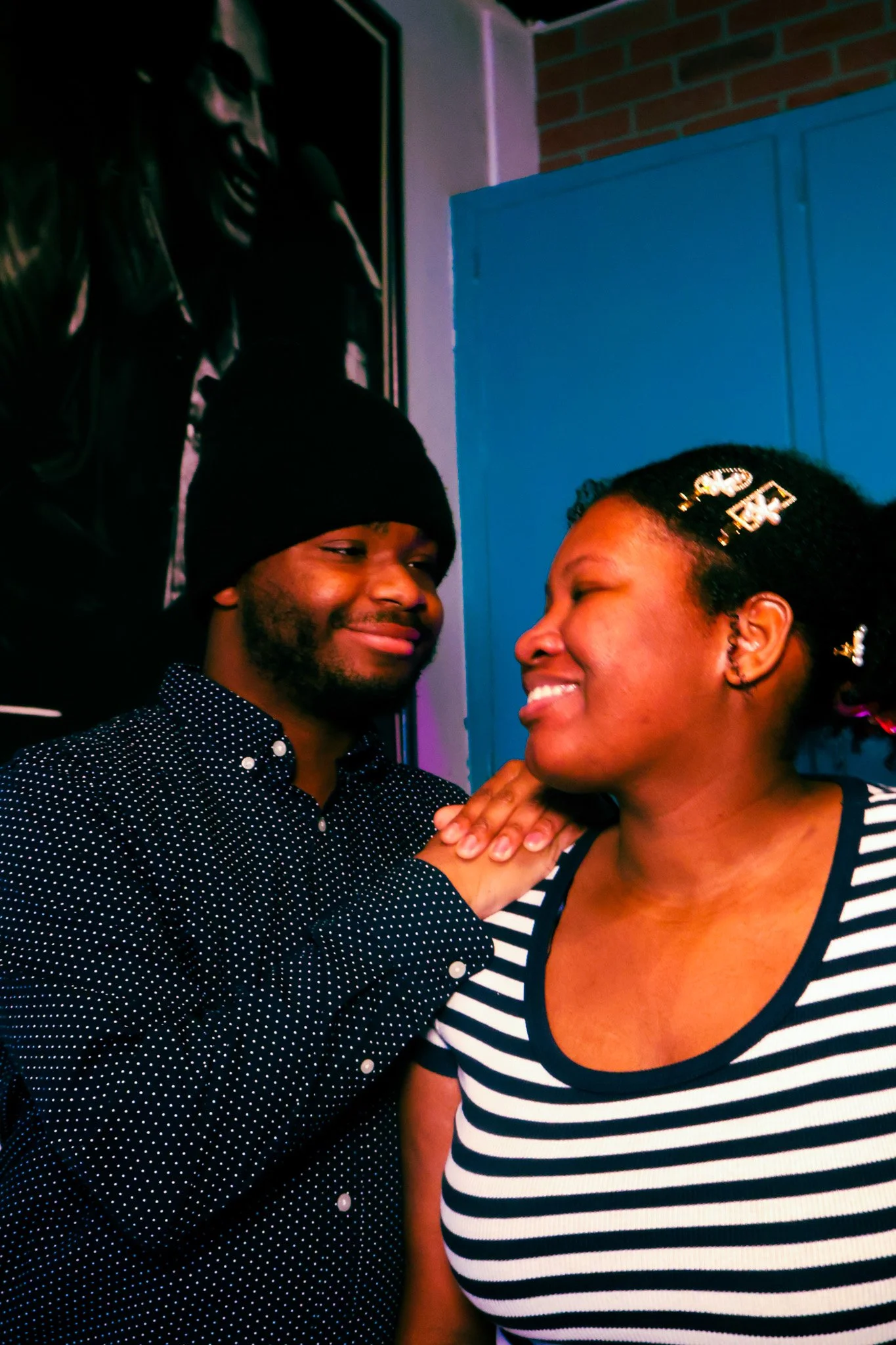 A man and woman sharing a close moment indoors, smiling and touching each other's shoulders, with a portrait on the wall behind them.