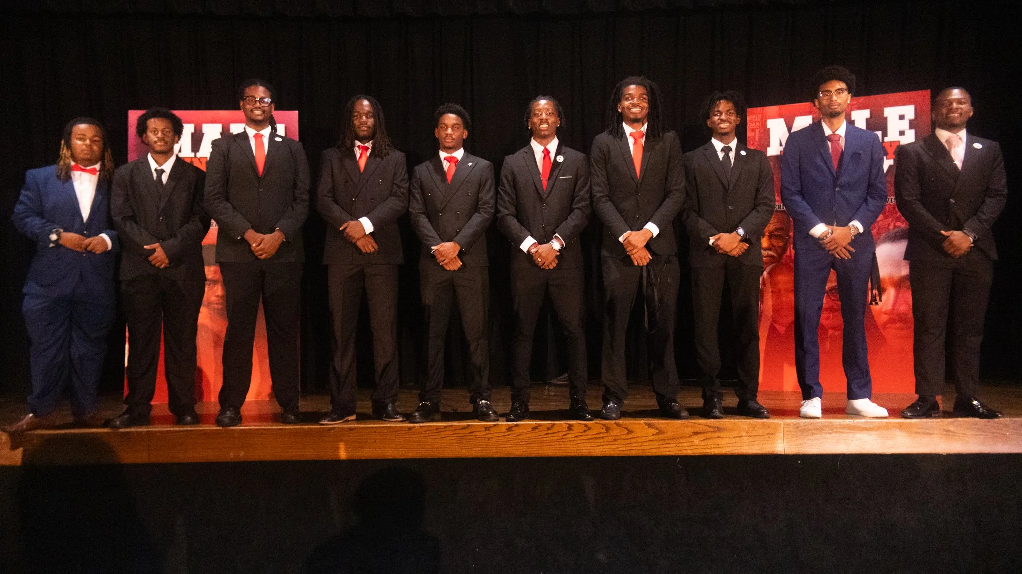 Group of eleven people dressed in formal suits standing on a stage in front of a black curtain with posters in the background.