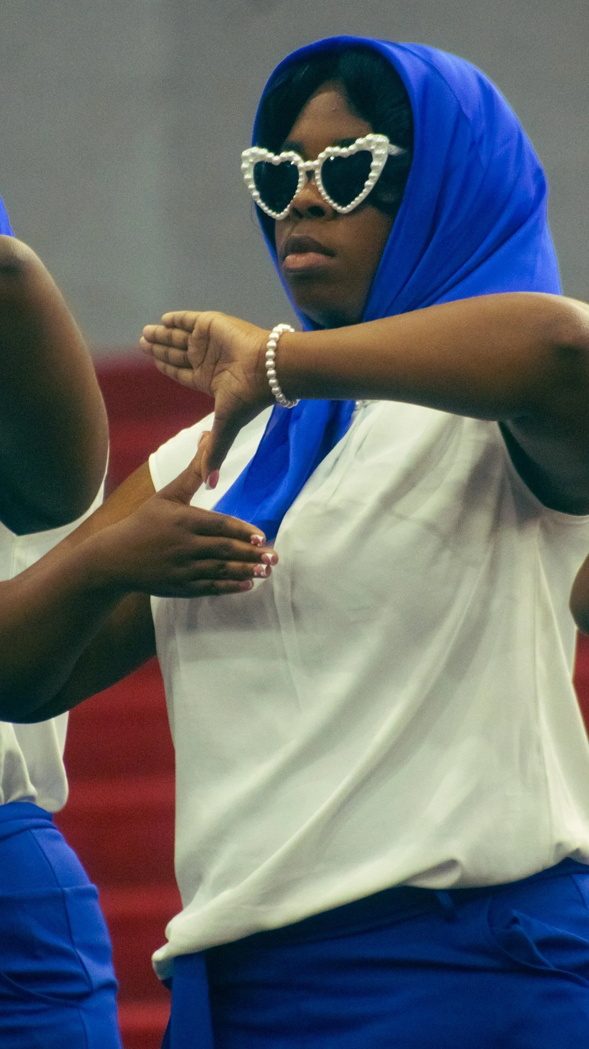 Person wearing pearl heart-shaped sunglasses, blue headscarf, white shirt, blue bottoms, and pearl bracelet, standing with arms crossed, with another person partially visible in front of them.