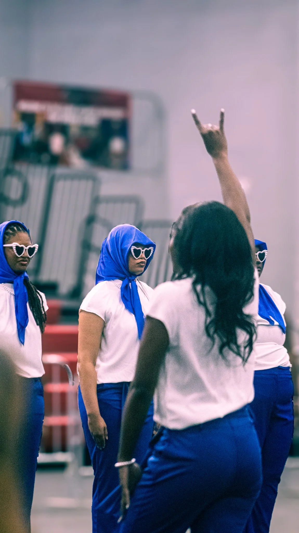 Group of women wearing matching white shirts, blue pants, blue headscarves, and large heart-shaped sunglasses inside a store, with one woman making a rock hand gesture.